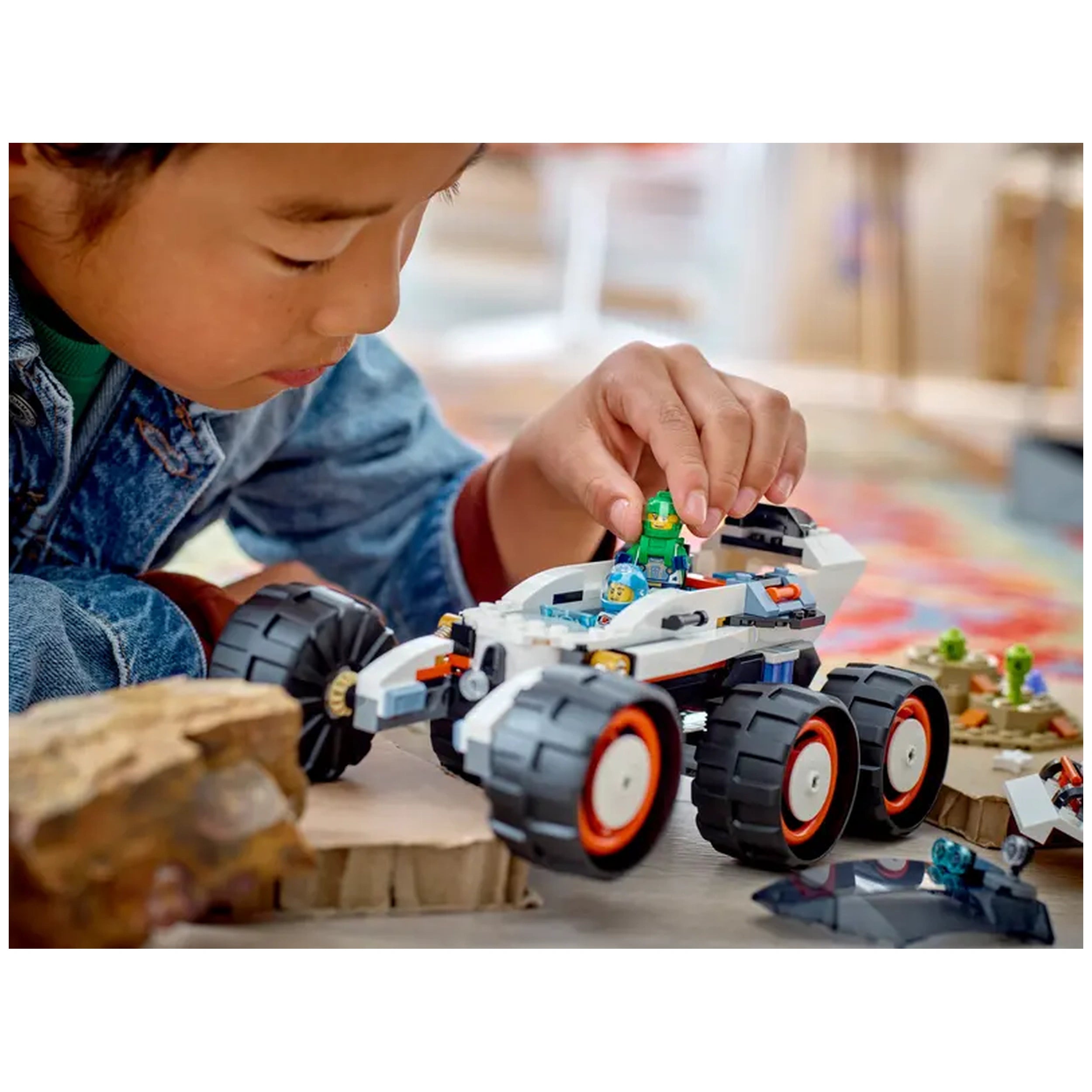 A child dressed in a denim jacket plays with a LEGO® City Space Explorer rover on a colorful rug, carefully placing a minifigure inside the vehicle. The rover features oversized wheels and a sleek design, surrounded by small alien figures and rocky terrain.