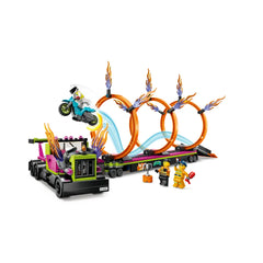 LEGO® City Stunt Truck with fiery exhaust stacks and a foldable ramp, featuring three orange rings of fire and a motorcycle mid-jump. Two Stuntz rider minifigures and a truck driver stand beside the truck, amidst tools and accessories for thrilling stunt challenges.