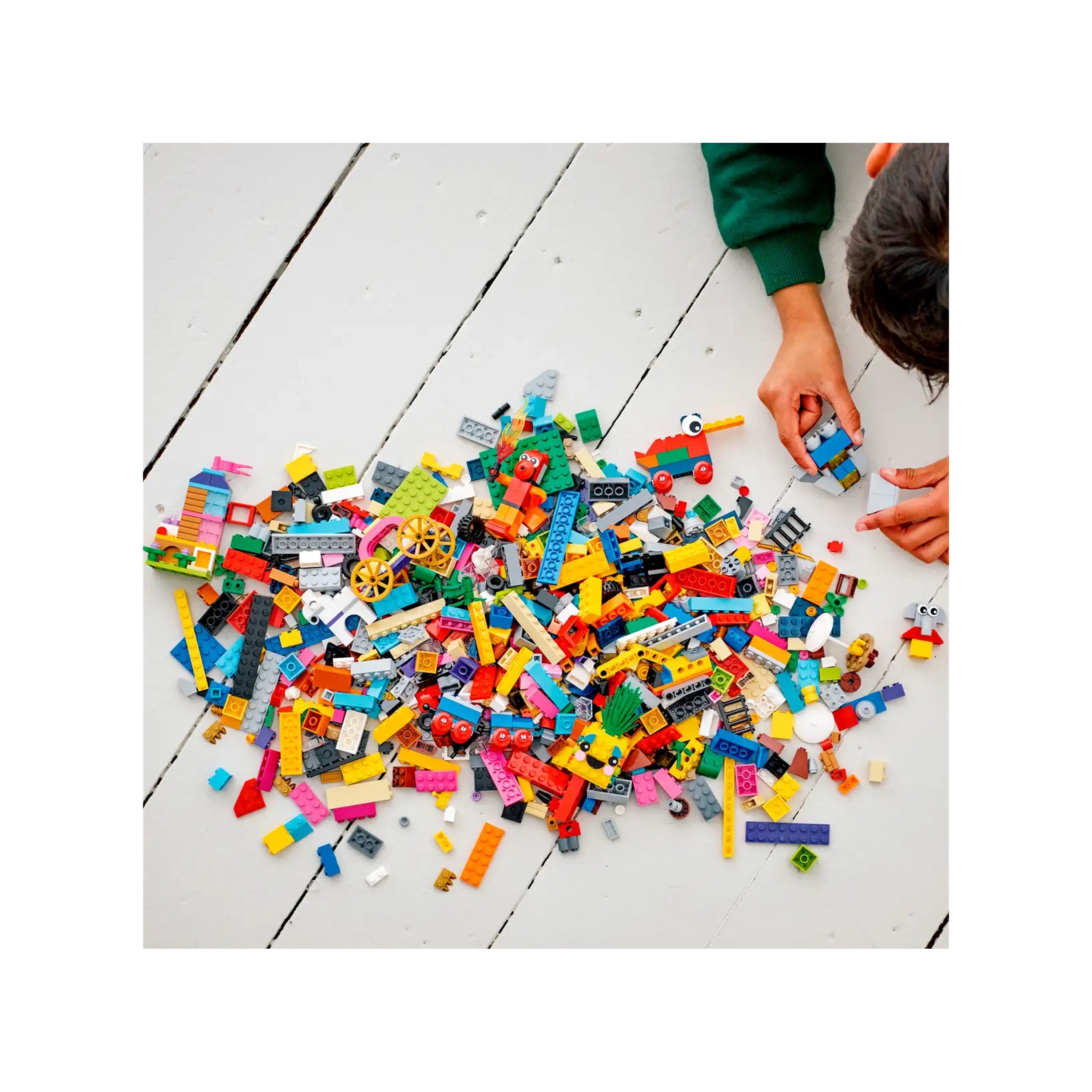 A colorful assortment of LEGO bricks scattered on a white wooden surface, with a child's hands building a mini toy.
