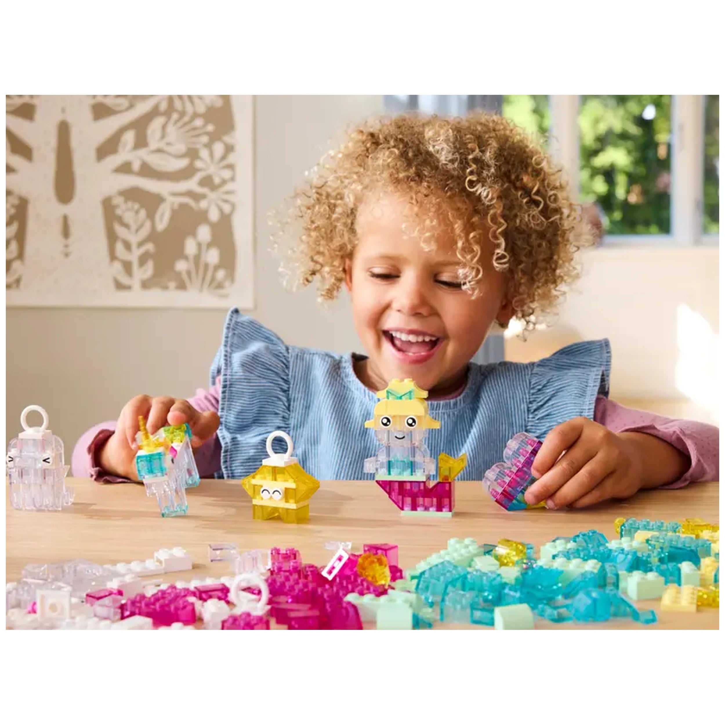 A joyful child with curly hair plays with colorful LEGO® transparent blocks, assembling whimsical models on a wooden table.