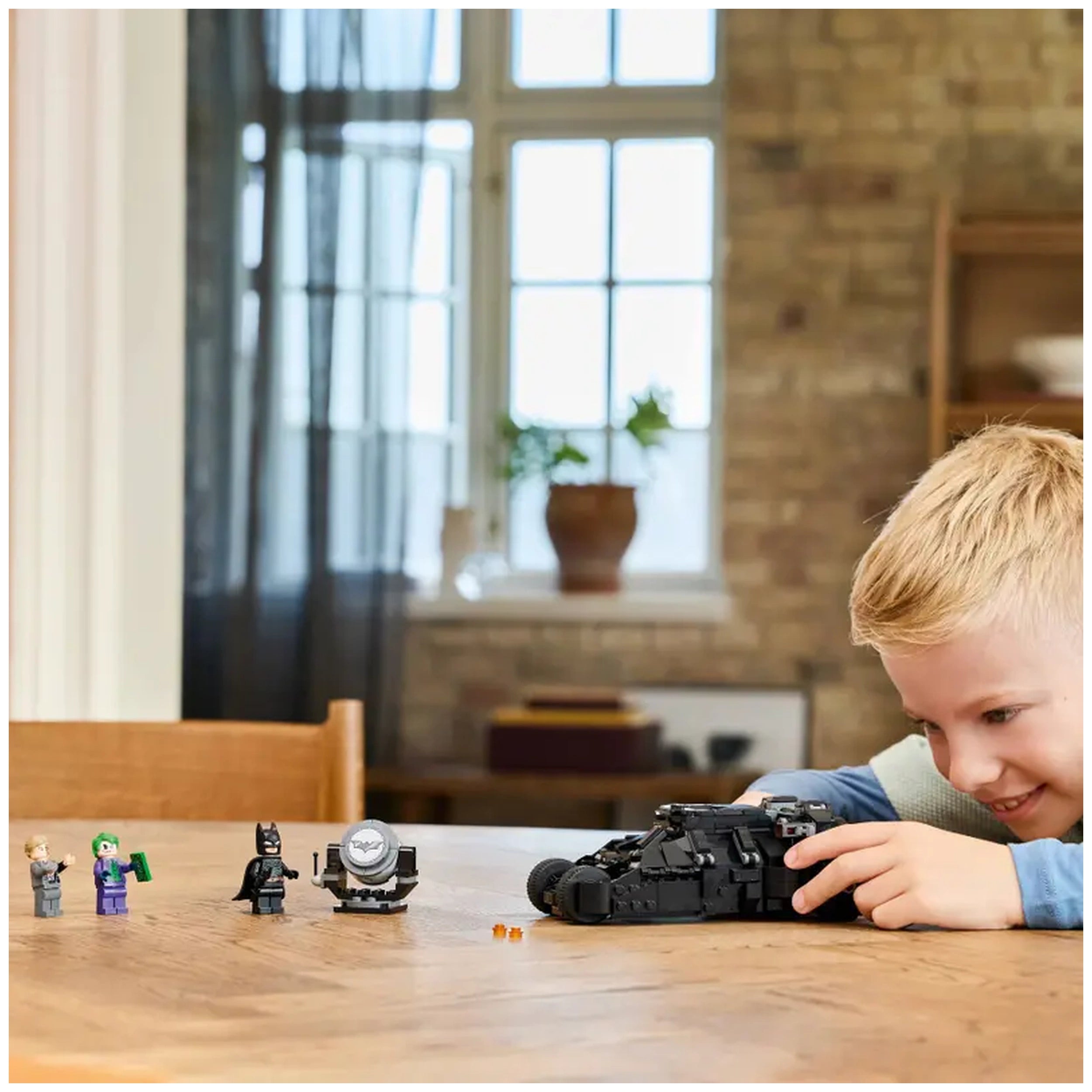A young boy with blonde hair smiles as he plays with the LEGO Batman Tumbler set on a wooden table, surrounded by superhero and villain figures, against a backdrop of bright windows and potted plants.