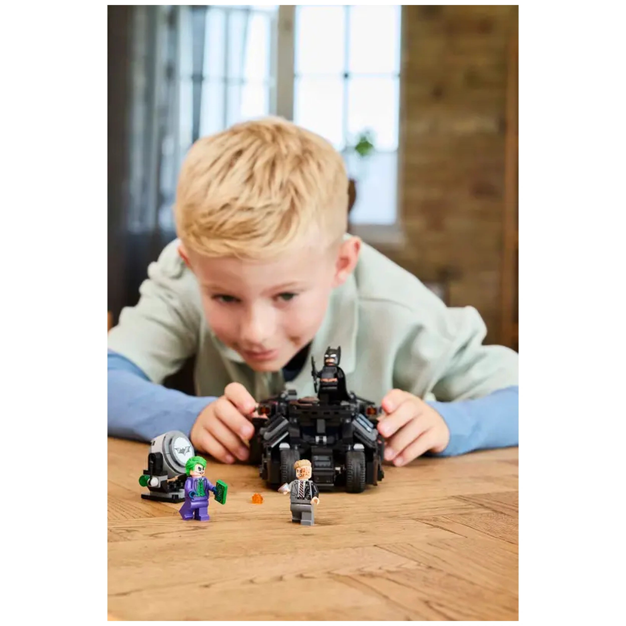 A child focuses intently on a LEGO Batman Tumbler set, featuring the Batmobile and minifigures of the Joker and Harvey Dent.