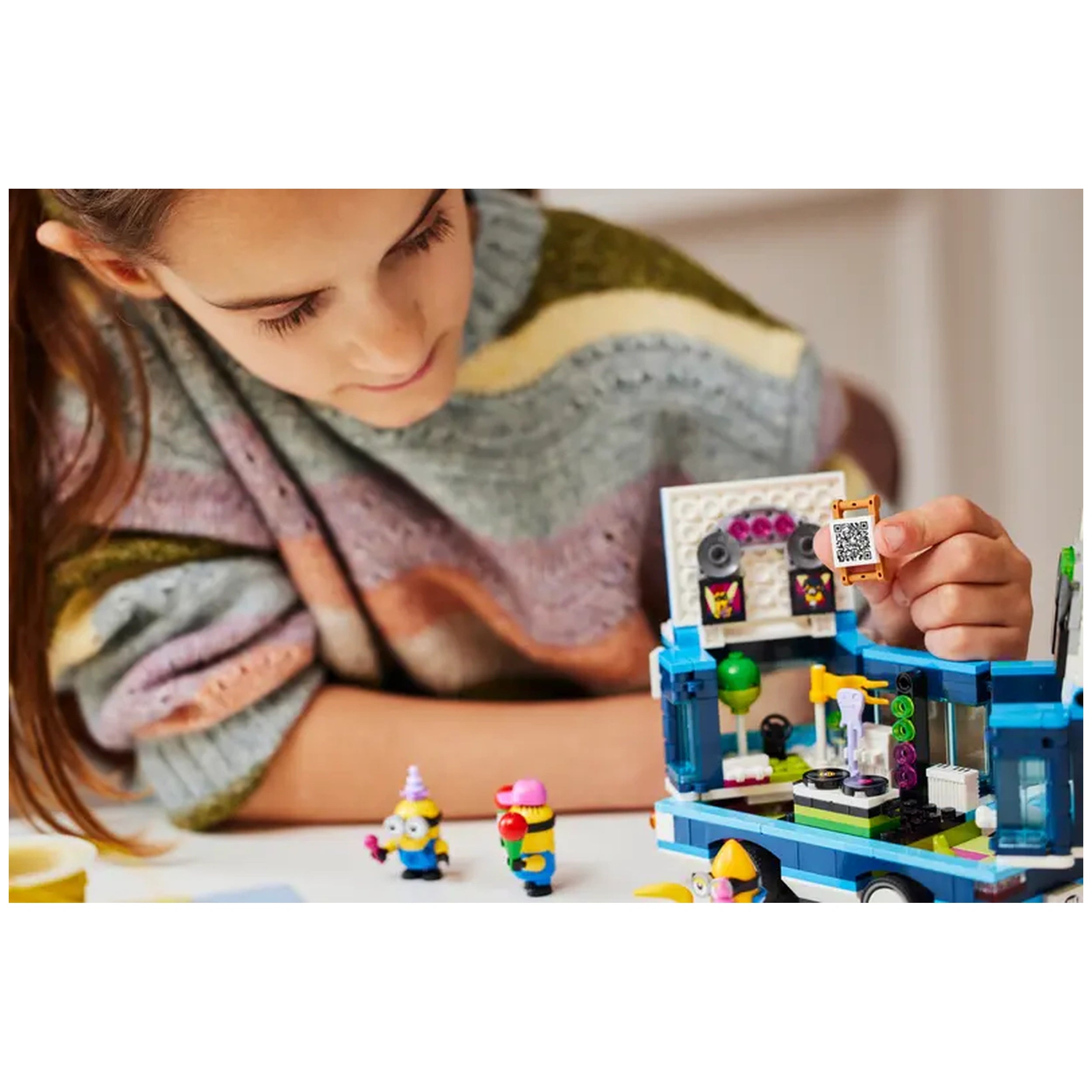 A child in a cozy sweater examines the LEGO® Minions' Music Party Bus set, holding a QR code piece. Two small Minion figures, one with a cone hat and another holding a red ball, are nearby. The colorful, vibrant bus features party decorations, creating an imaginative scene.