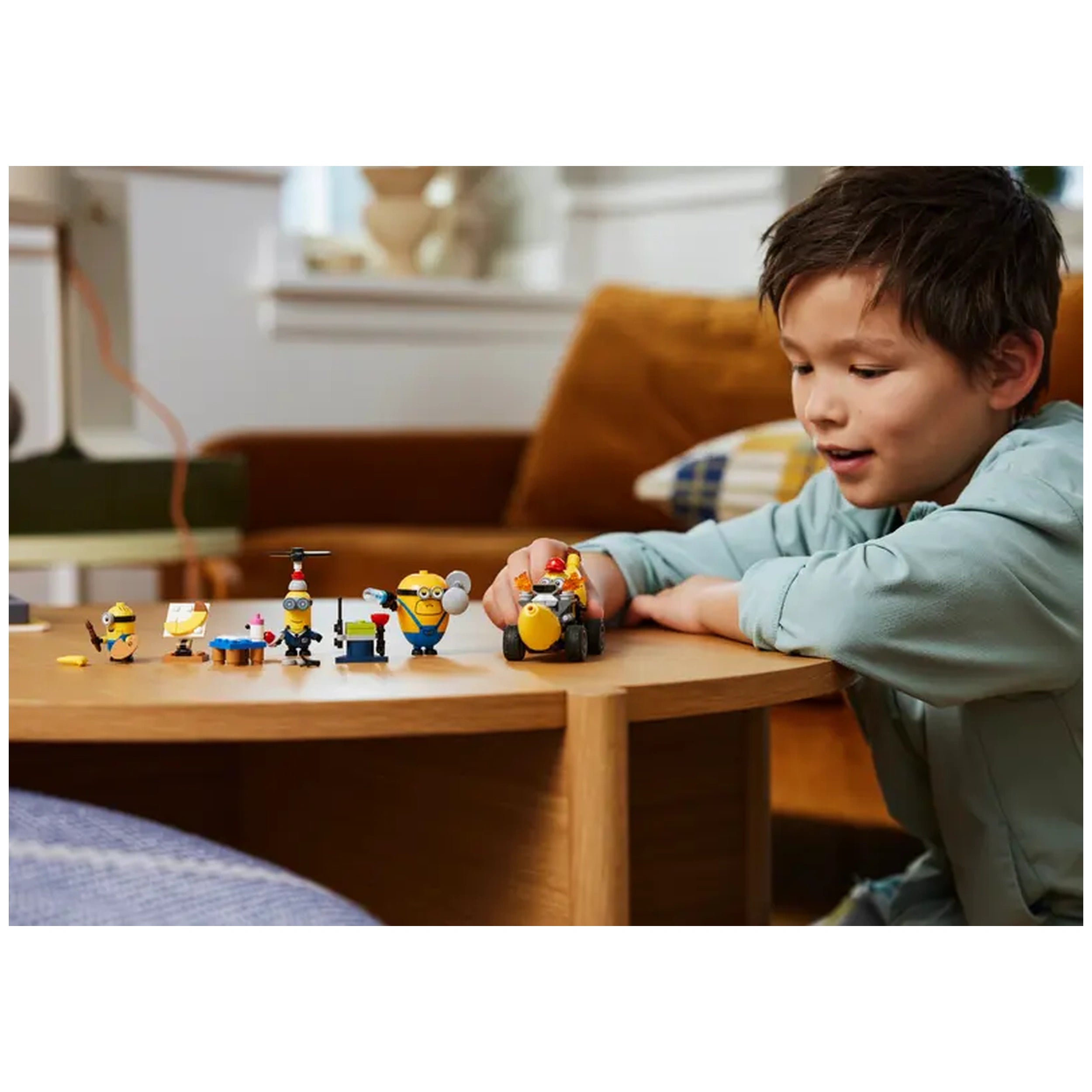 A young boy smiles while playing with LEGO® Minion figures and the Banana Car set on a wooden table, surrounded by colorful toys.