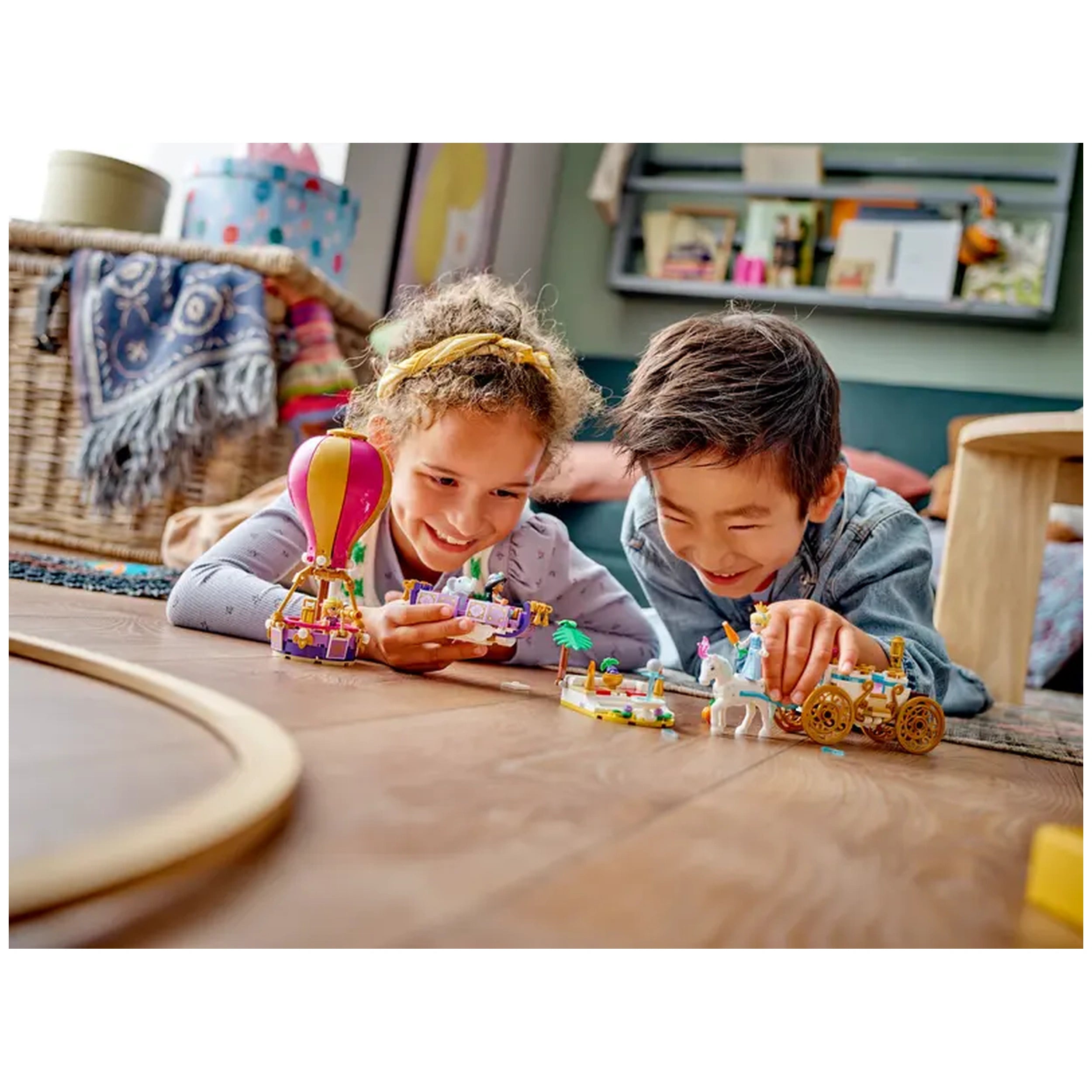 Two children play on the floor, joyfully interacting with LEGO Disney Princess sets. A colorful hot-air balloon and carriages are visible, surrounded by playful mini-doll figures and a unicorn, enhancing their imaginative adventure.