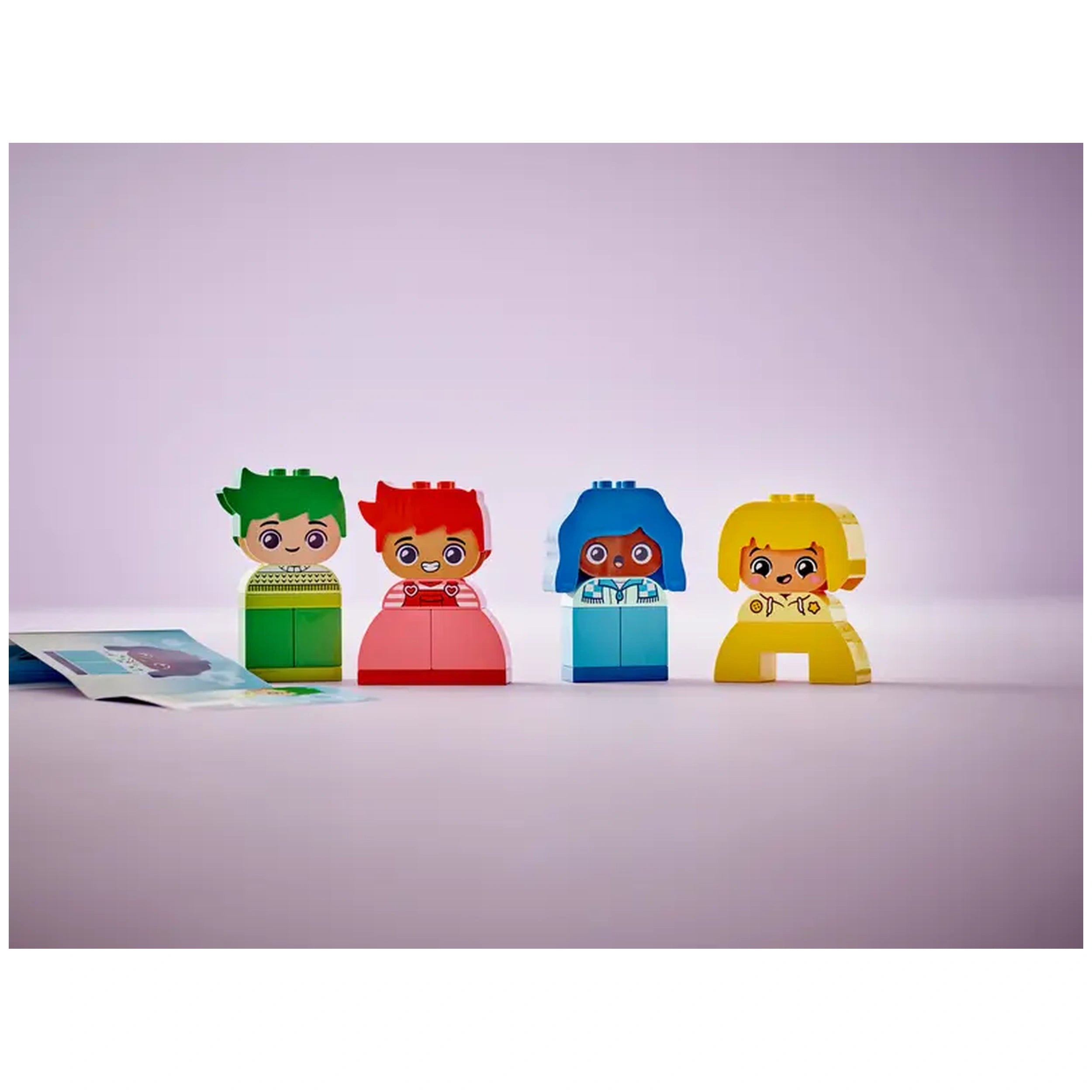 Four vibrant LEGO® DUPLO® characters stand against a soft pink backdrop, showcasing interchangeable facial expressions. The characters include a green-haired boy in a patterned sweater, a red-haired girl, a blue-haired child surprised by emotions, and a cheerful yellow figure. To the left, a colorful instruction booklet lies partially visible, enhancing the playful scene.
