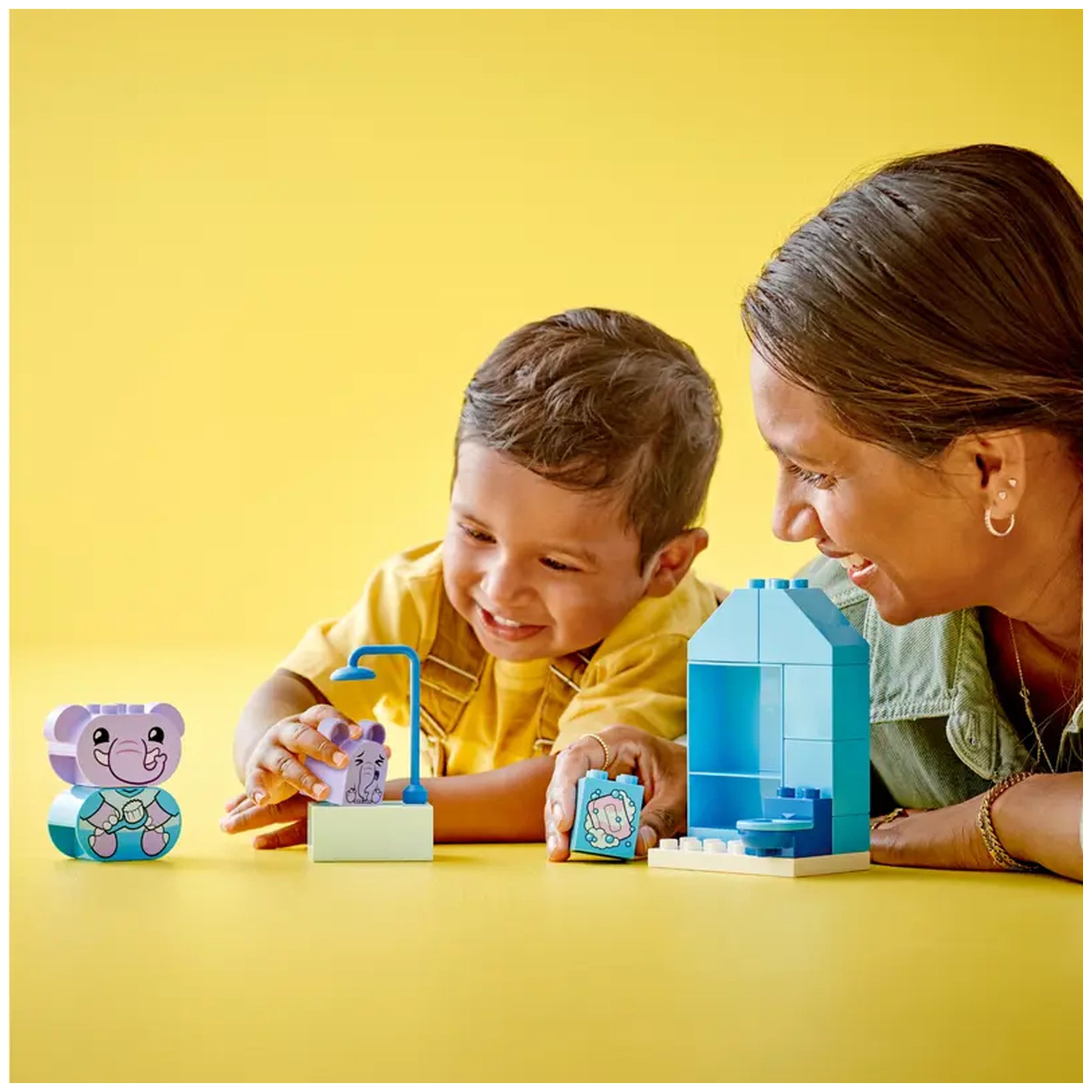 A cheerful child in a yellow shirt plays with LEGO® DUPLO® bath time toys, including an elephant figure and a colorful bathroom set on a bright yellow background. An adult nearby smiles, engaging in imaginative play.