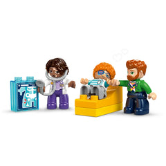LEGO® DUPLO® figures in a doctor’s office scene, including a doctor with a stethoscope, a child patient on a yellow examination bed, and a parent holding the child's hand, with an X-ray prop nearby.