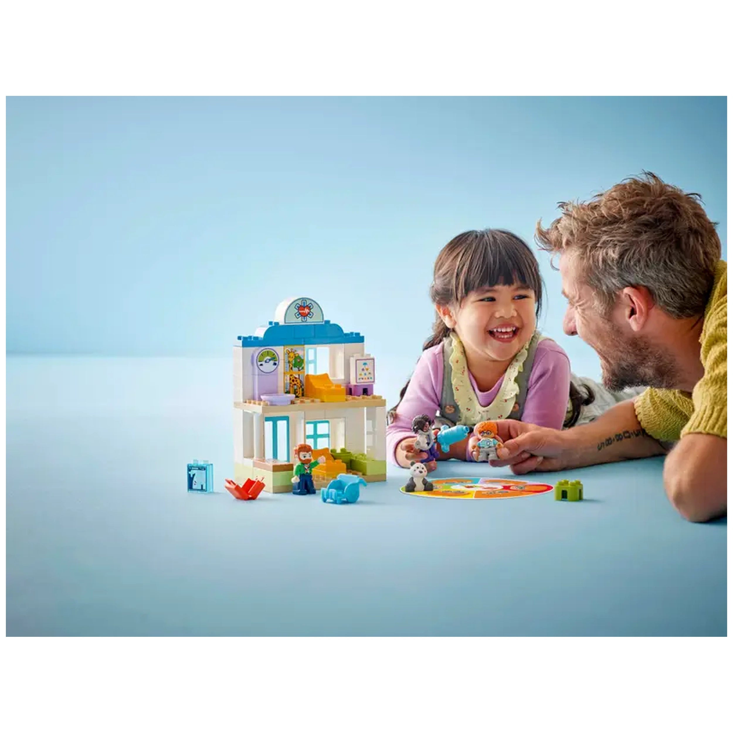A joyful child and smiling adult play with the LEGO® DUPLO® Town hospital set, featuring figures and colorful props, on a light blue background.