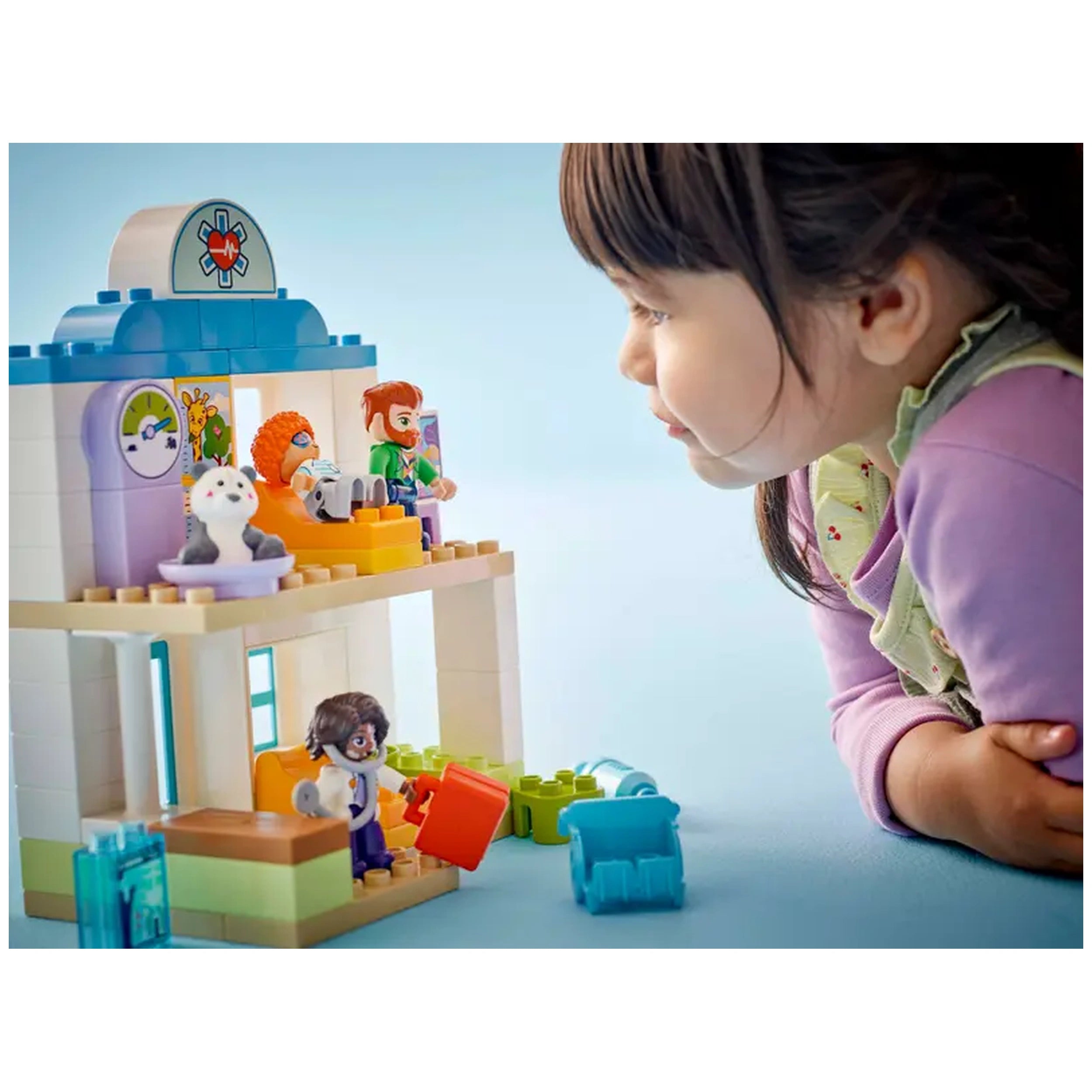 A toddler gazes intently at a colorful LEGO® DUPLO® hospital playset, featuring a doctor, child, and a teddy bear.