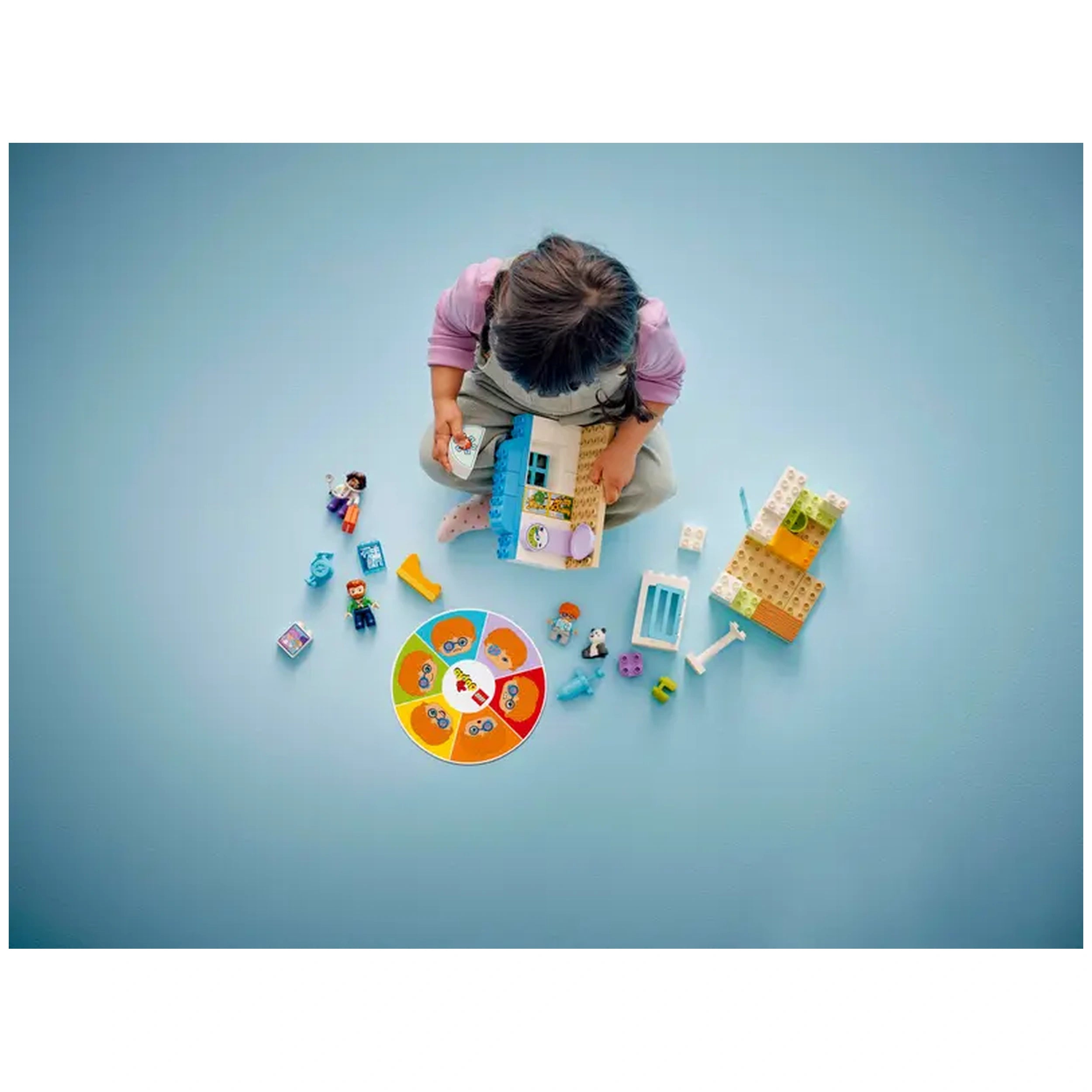 A toddler with dark hair sits on a blue surface, holding a LEGO® DUPLO® playset piece. Surrounding them are colorful building blocks, character figures, and a circular gameboard, inviting imaginative play in a hospital-themed setting.