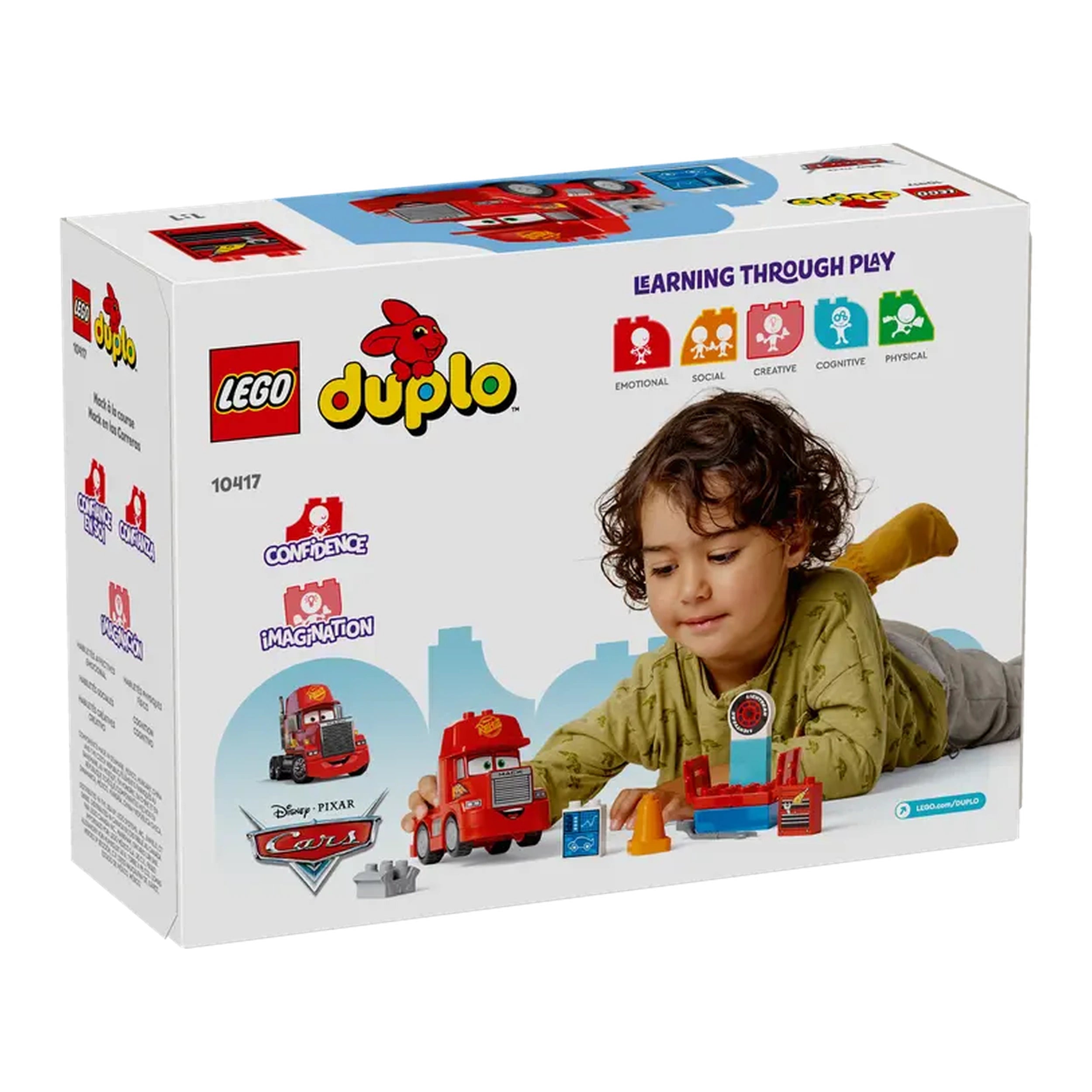 Young child with brown curly hair, playing with a LEGO® DUPLO® Mack at the Race construction set. The vibrant red Mack toy is near a blue tuning station, alongside colorful accessories, on a white box featuring the DUPLO logo.