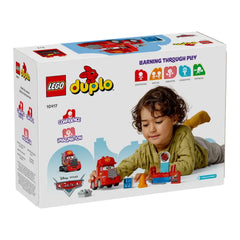 Young child with brown curly hair, playing with a LEGO® DUPLO® Mack at the Race construction set. The vibrant red Mack toy is near a blue tuning station, alongside colorful accessories, on a white box featuring the DUPLO logo.