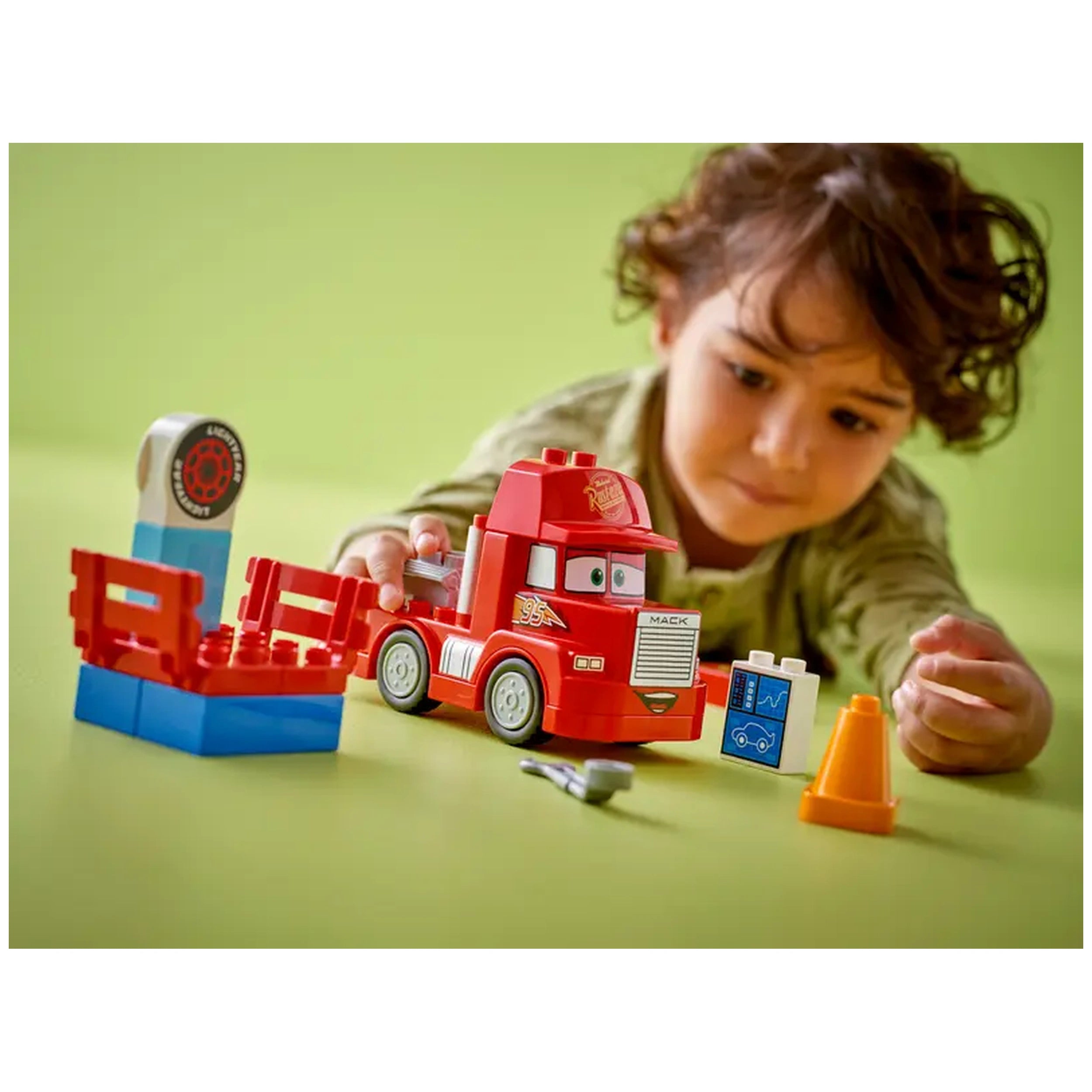 A young child interacts with a bright red LEGO® DUPLO® Mack truck, surrounded by colorful accessories on a green background. The child focuses intently, reaching for a small blue accessory resembling a car monitor. Nearby, a tuning station and an orange traffic cone add to the imaginative play, enhancing the excitement of the Disney and Pixar Cars-themed set.