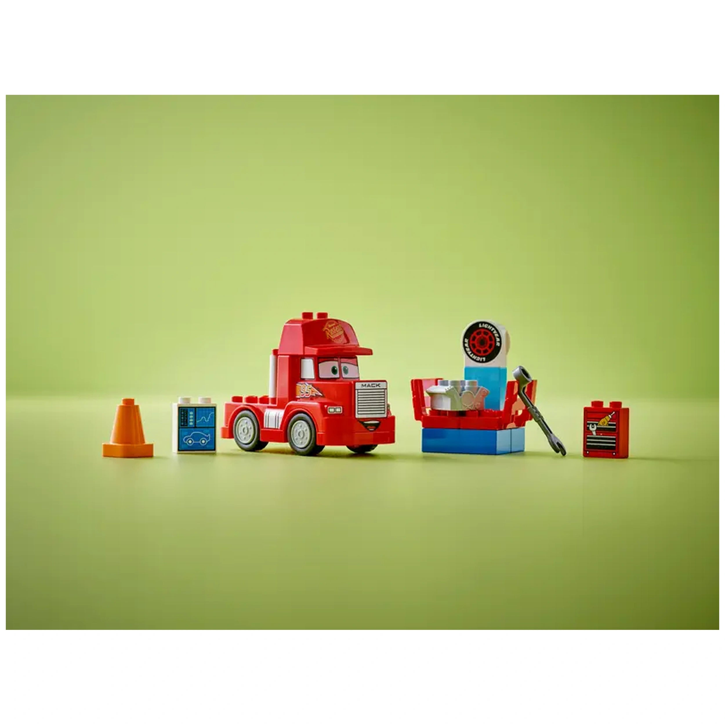 Red LEGO® DUPLO® Mack truck with smiling face, next to a blue tuning station, orange cone, engine brick, and wrench on a light green background.
