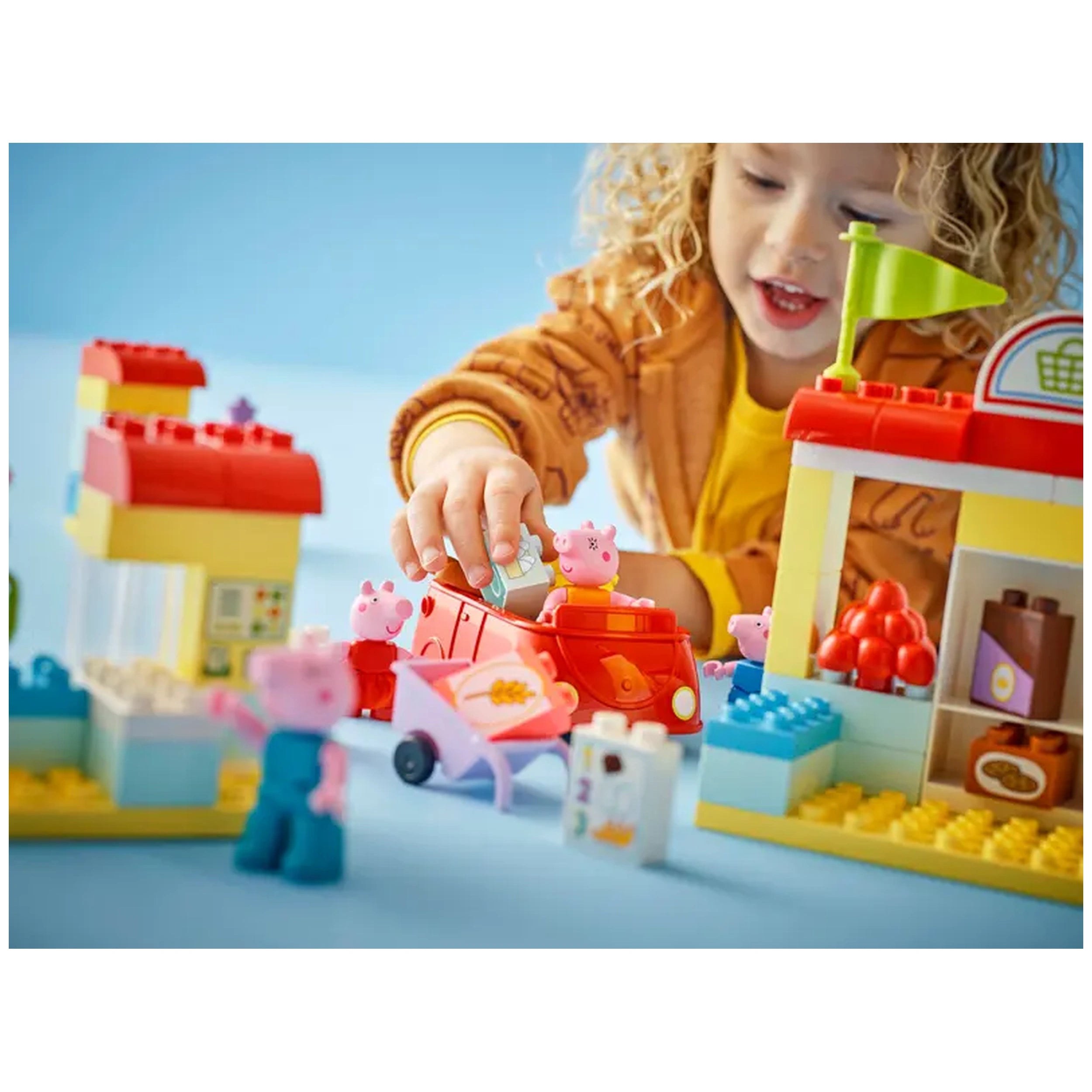 A child joyfully plays with the LEGO® DUPLO® Peppa Pig Supermarket set, placing Peppa Pig figures in a red toy car near a colorful grocery store with a green flag.