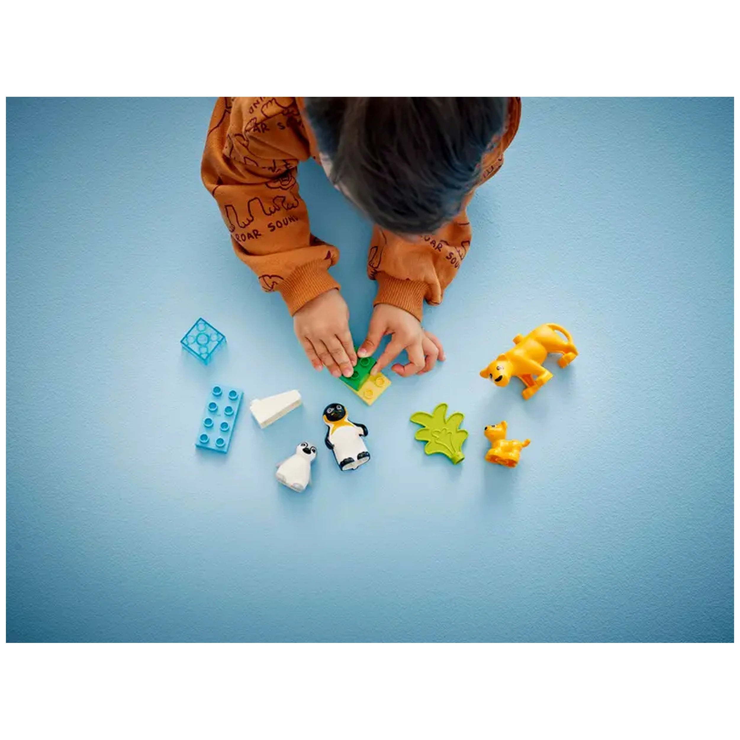 Young child in a brown sweater assembling green LEGO® DUPLO® bricks, surrounded by colorful animal figures including penguins and a lion on a light blue surface.