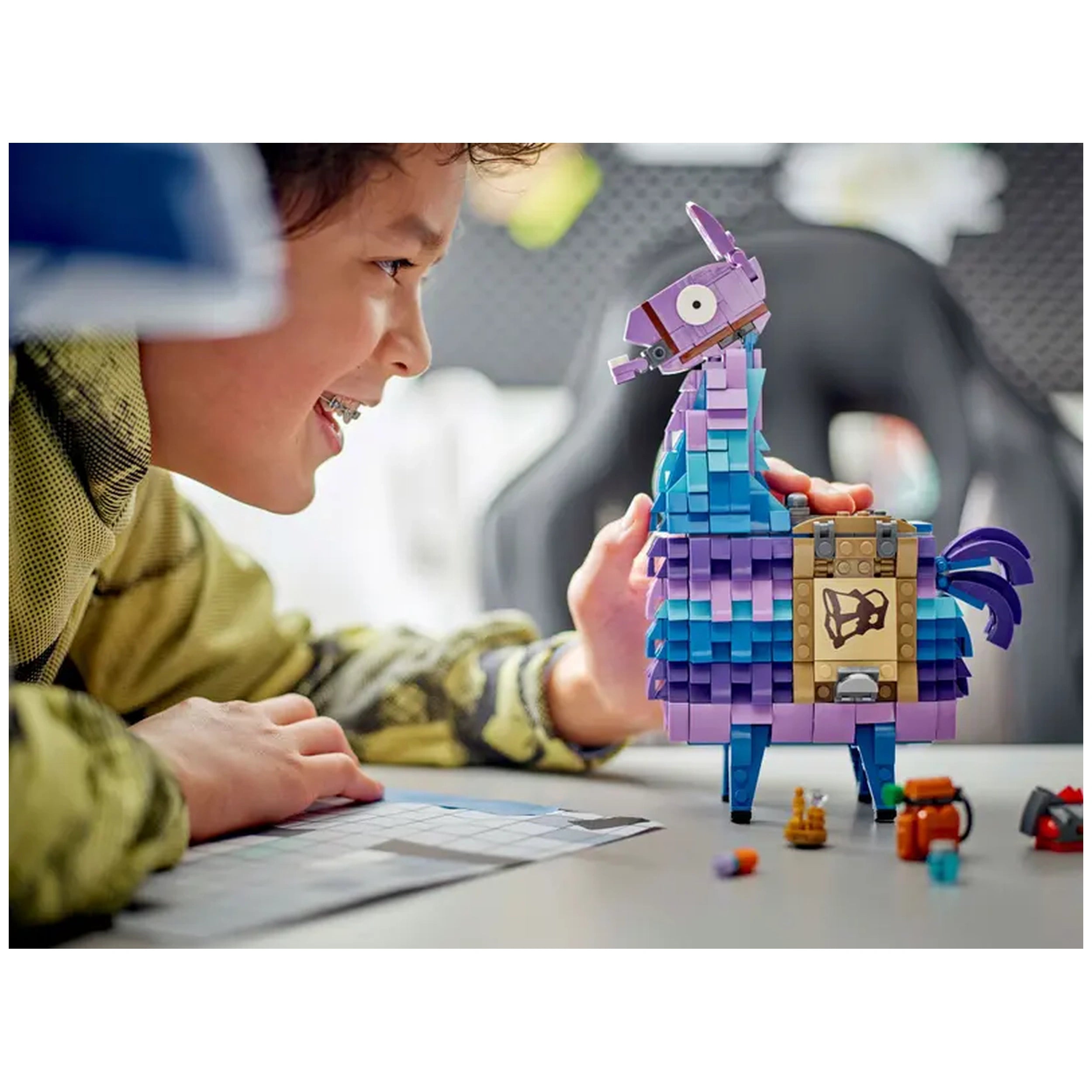 A cheerful boy interacts with a vibrant LEGO Fortnite Supply Llama, showcasing its colorful design with details like a movable head. On the table, scattered accessories like a grappler and slurp juice complement the play scene.