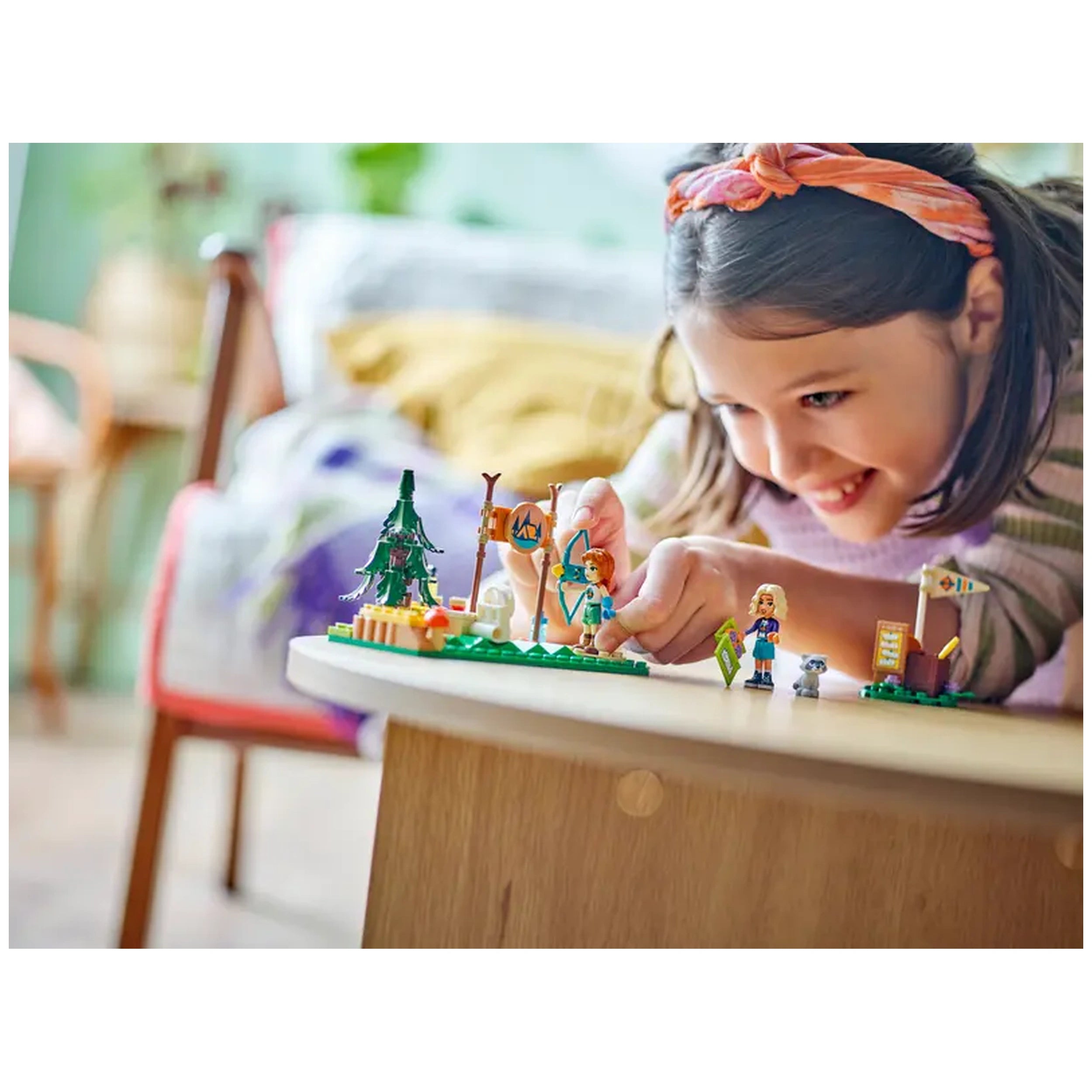 A young girl joyfully plays with the LEGO® Friends Adventure Camp Archery Range set on a wooden table, engaging with mini-dolls and creative accessories.