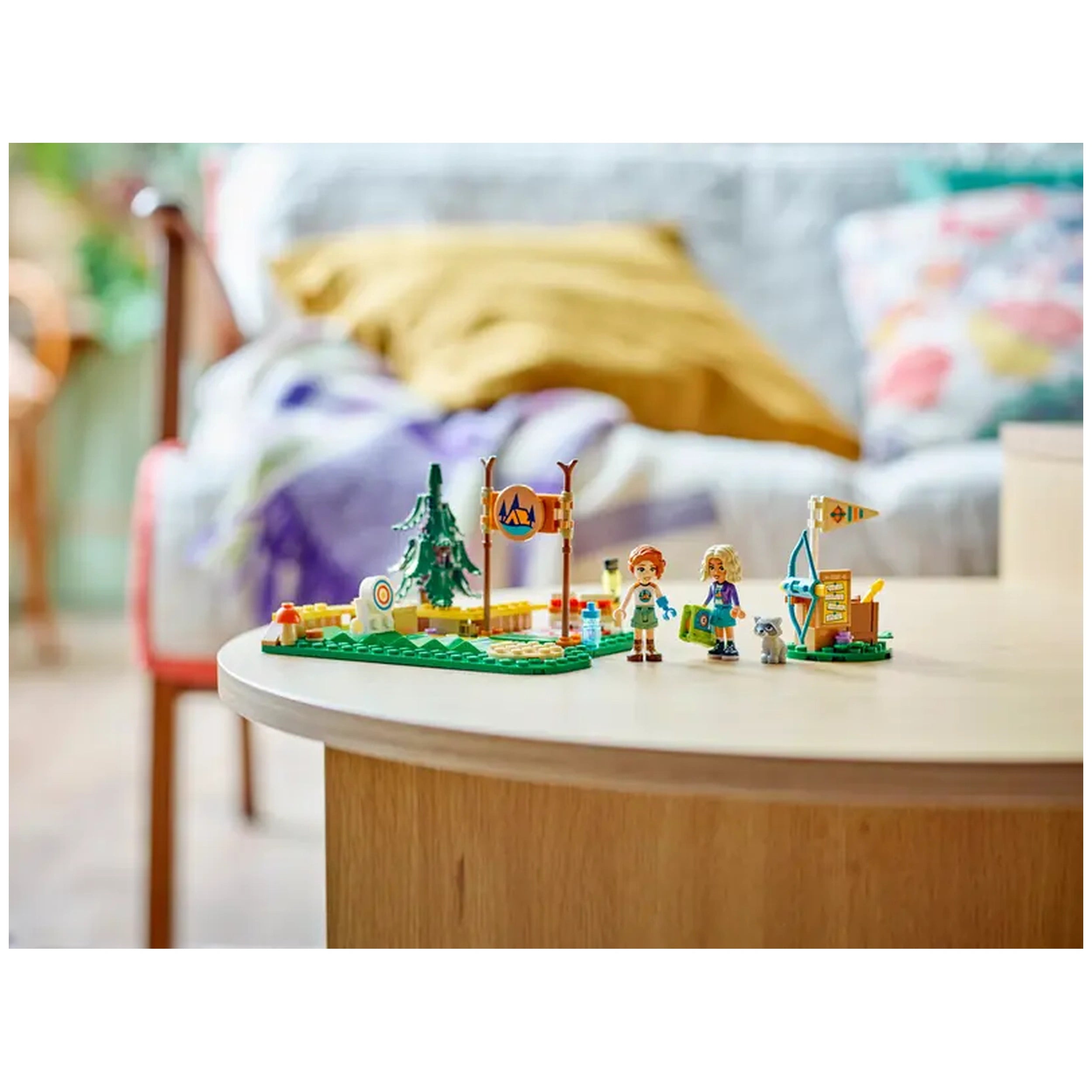Two mini-dolls, Autumn and Nova, stand on a green LEGO base near an archery range, complete with a tree, target, and playful raccoon figure. A colorful camping sign and flag enhance the vibrant outdoor scene.