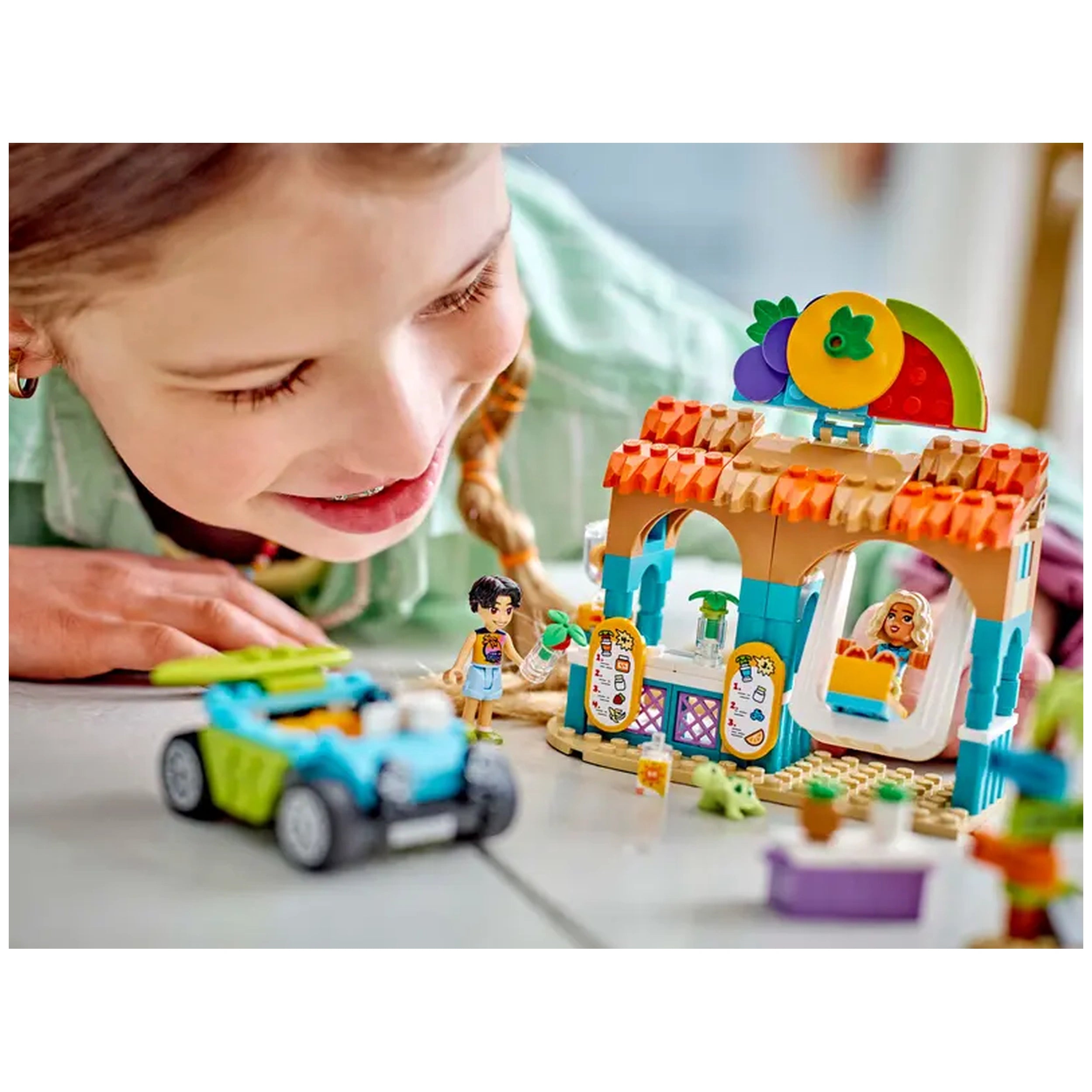 A child plays with the LEGO® Friends Beach Smoothie Stand, featuring a colorful smoothie stand and two mini-dolls, surrounded by a beach buggy.