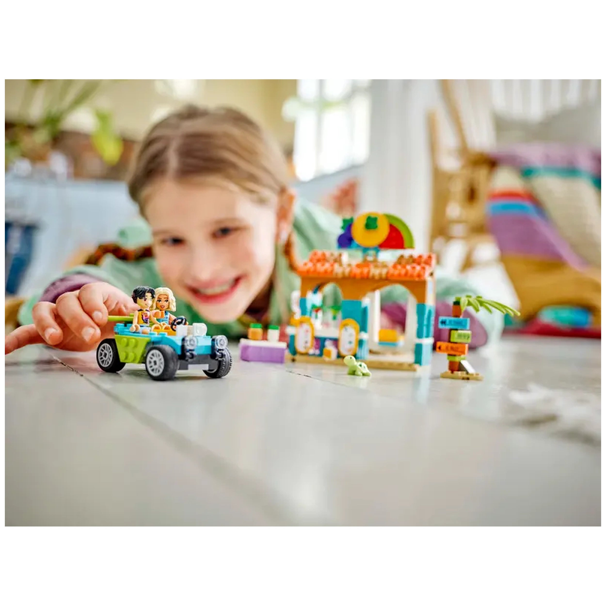 A smiling child plays with a LEGO® Friends Beach Smoothie Stand set, featuring a colorful smoothie stand and a beach buggy.