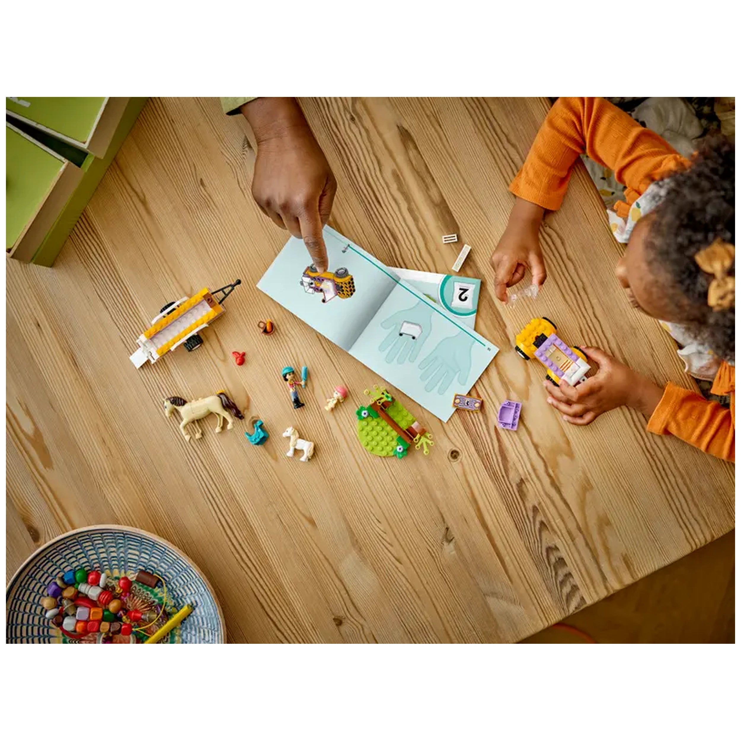 A child's hand reaches for a LEGO® Friends construction set on a wooden table, featuring figures, a horse, and a pony trailer. An adult's hand points at the instruction booklet with step two illustrated, while colorful building pieces and a basket of assorted toys scatter nearby.
