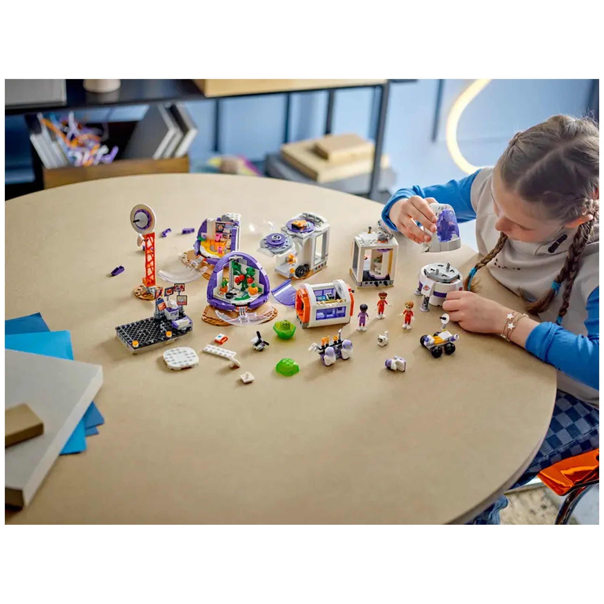 A child interacts with the LEGO® Friends Mars Space Base set on a round table, surrounded by vibrant mini-dolls, a rocket, and various buildable elements, fostering imaginative play.