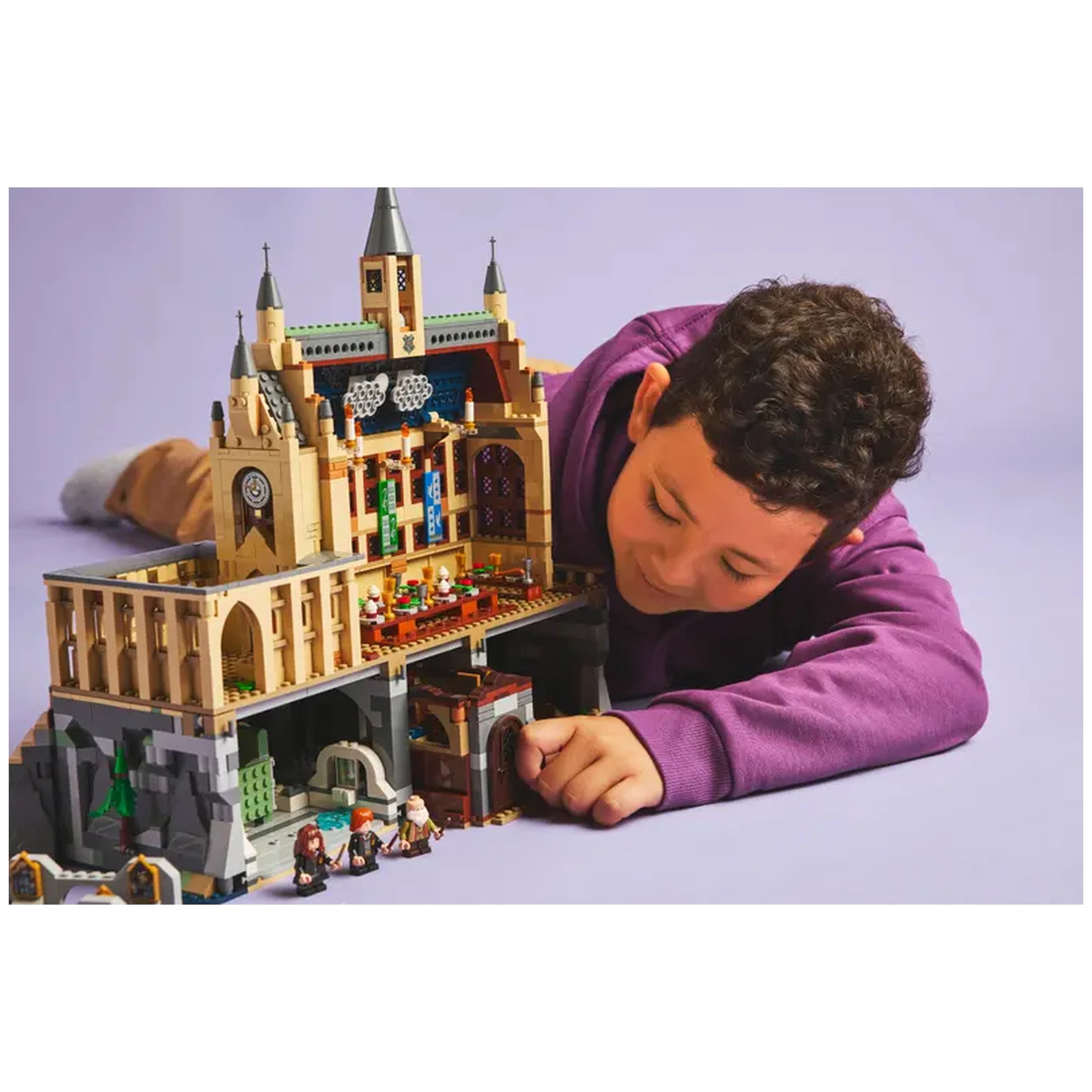 A child in a purple sweater is engaged with a detailed LEGO Hogwarts Castle set, showcasing vibrant classroom scenes and playful characters on a light purple backdrop.