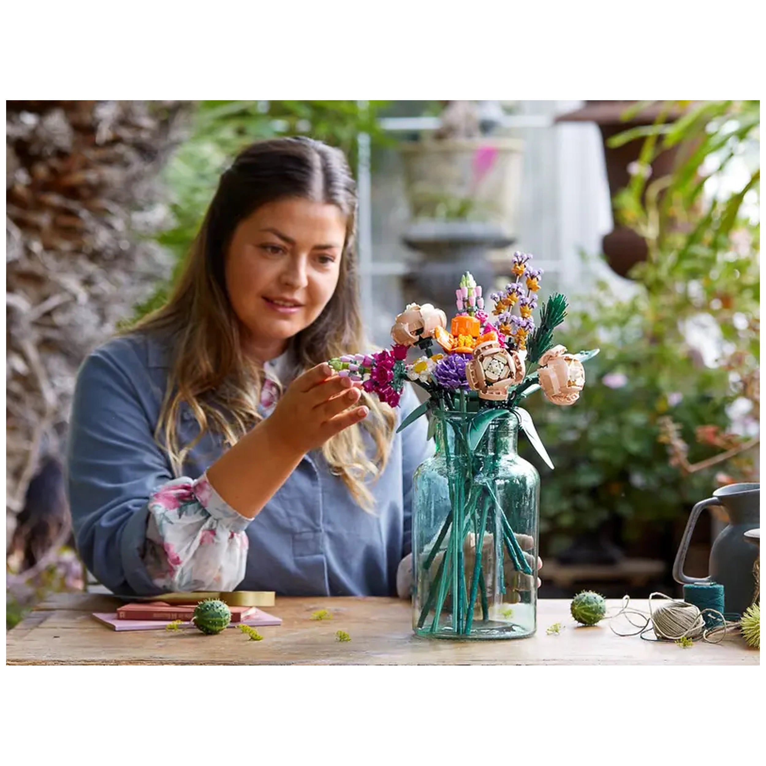 A woman in a blue shirt carefully arranges a vibrant LEGO® Flower Bouquet in a glass vase on a wooden table, surrounded by greenery.