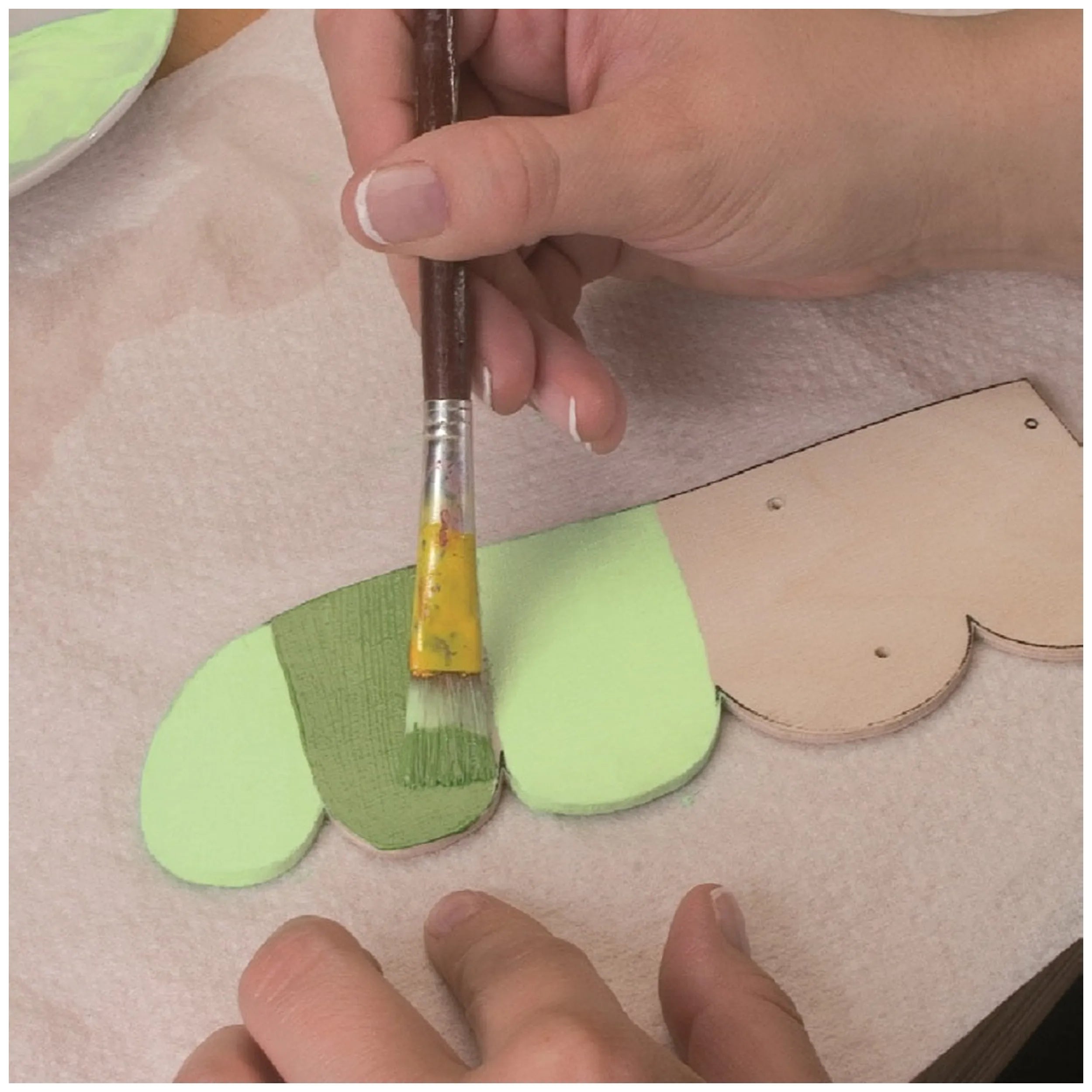 Hands painting wooden cutouts with green paint from the Prebaro Fretwork Kit, emphasizing creativity and craftsmanship.