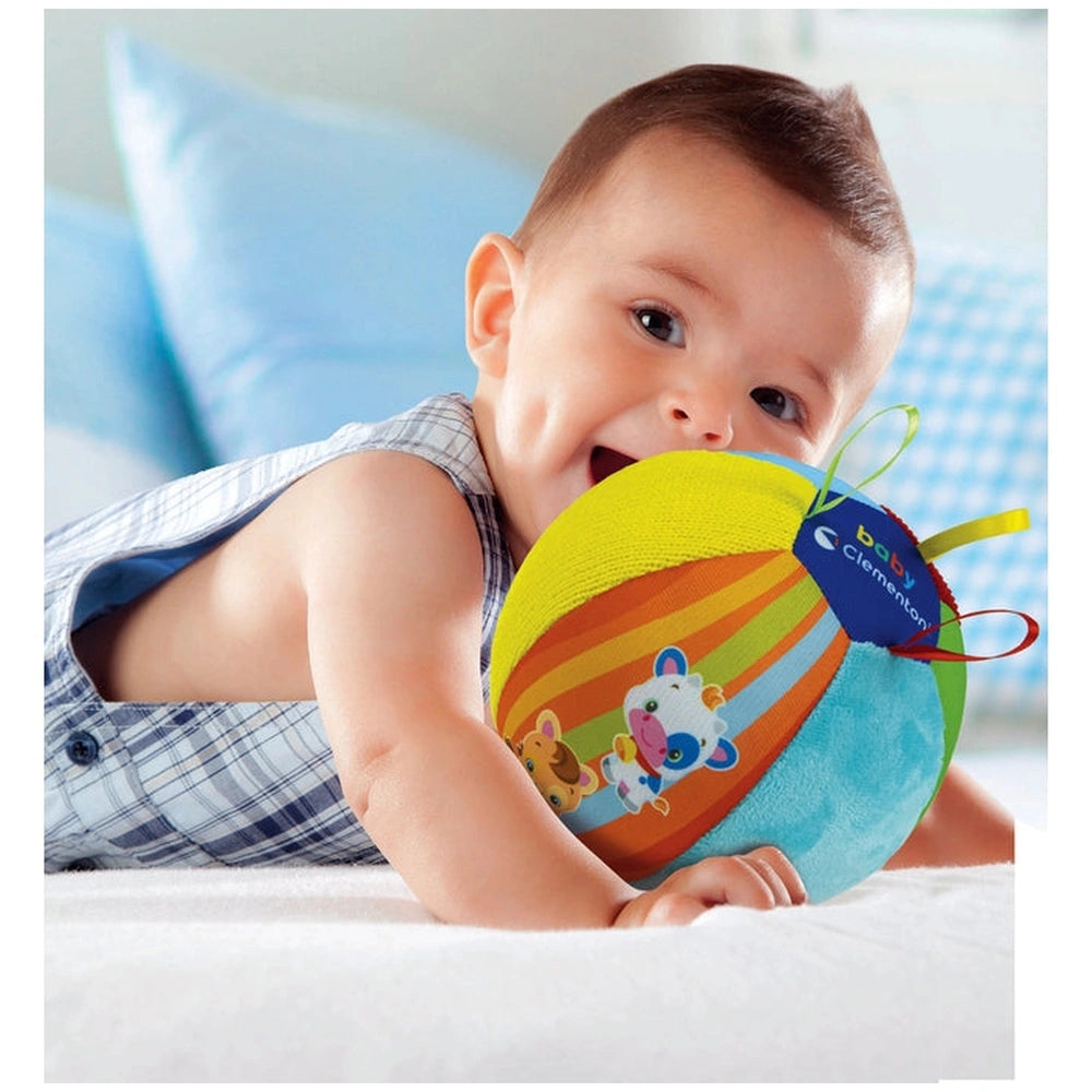 Baby smiles while holding a colorful plush toy on a white bed with a blue pillow, wearing a plaid shirt.