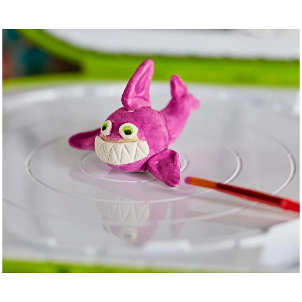 Plush pink shark toy with white underside, green eyes, and purple fin on a smooth surface in the CRAYOLA Clay Sculpting Stati