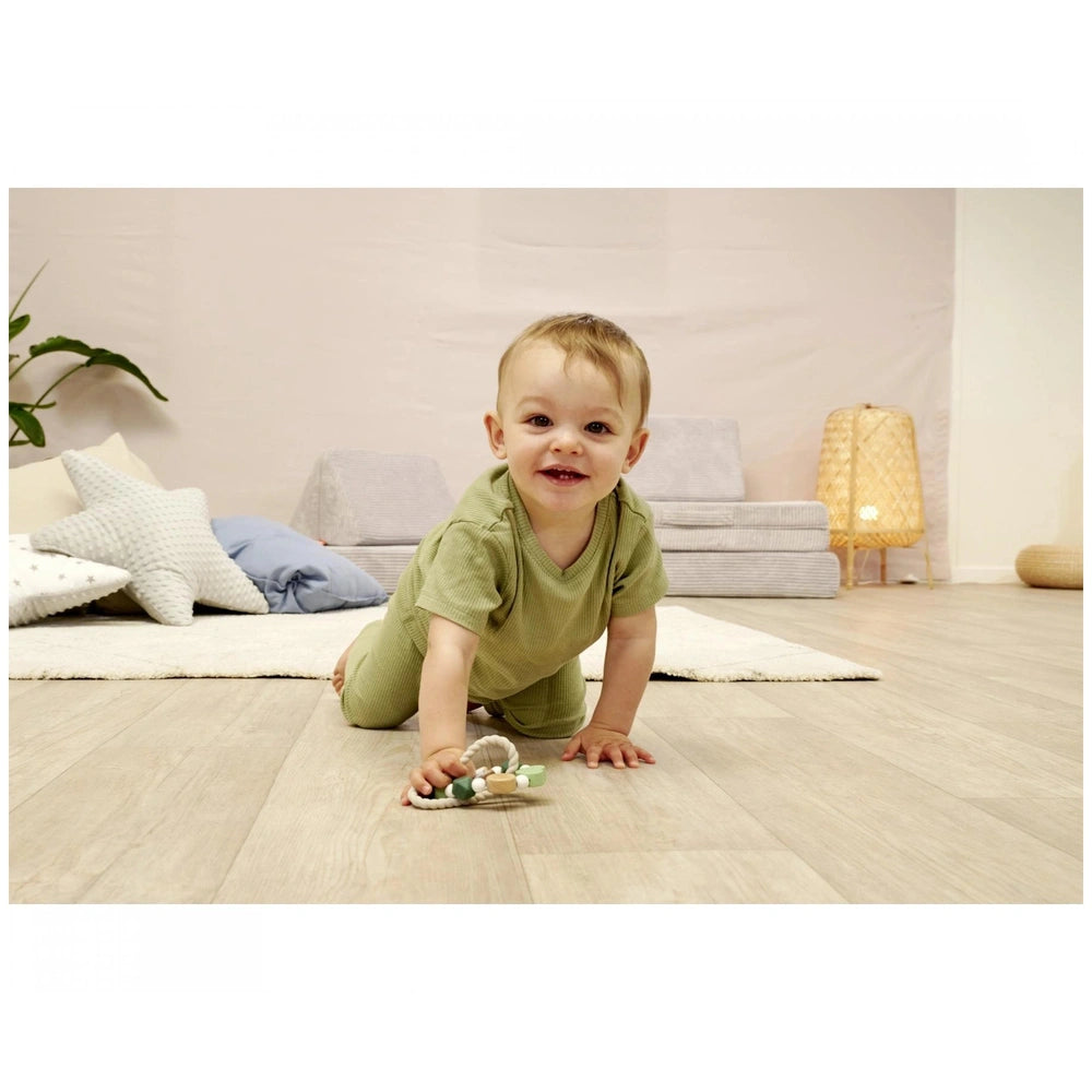 Baby sitting on a light wooden floor, smiling in a green outfit, surrounded by a soft pastel nursery setting.