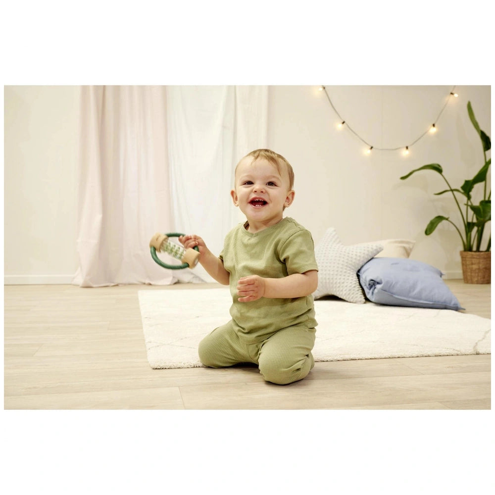 Eichhorn Baby HiPP Sound Rattle is held by a smiling child in a green outfit, sitting on a white rug in a bright room.