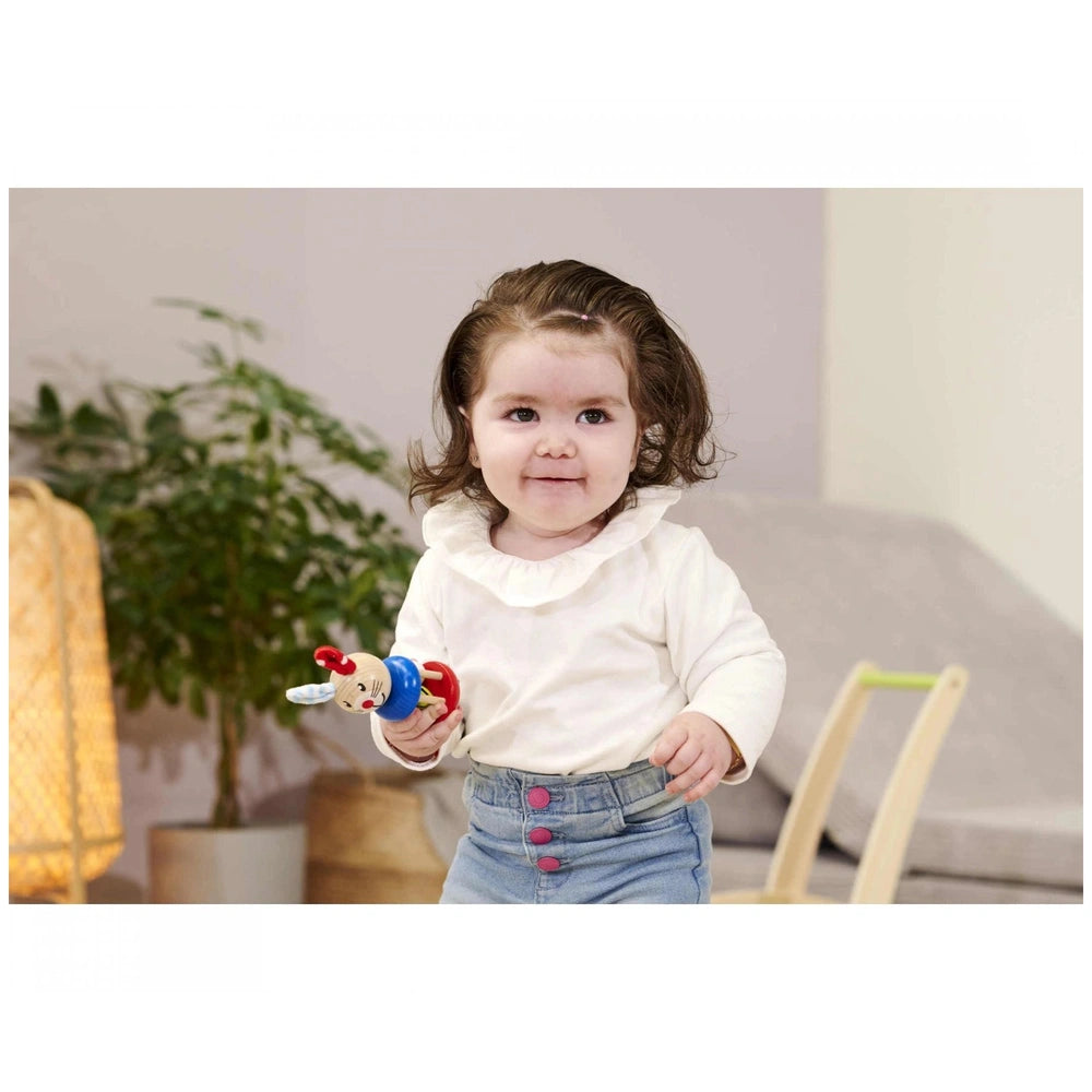Eichhorn Baby Wooden Rattle Rabbit is held by a smiling toddler indoors, surrounded by a couch and a plant.