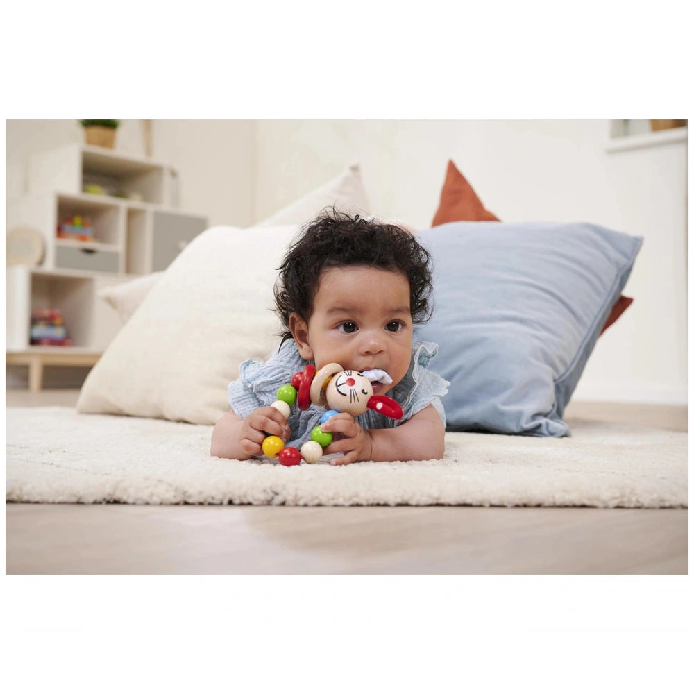 Eichhorn baby wooden teething ring rabbit held by a young child sitting on a soft surface with a stuffed toy nearby.