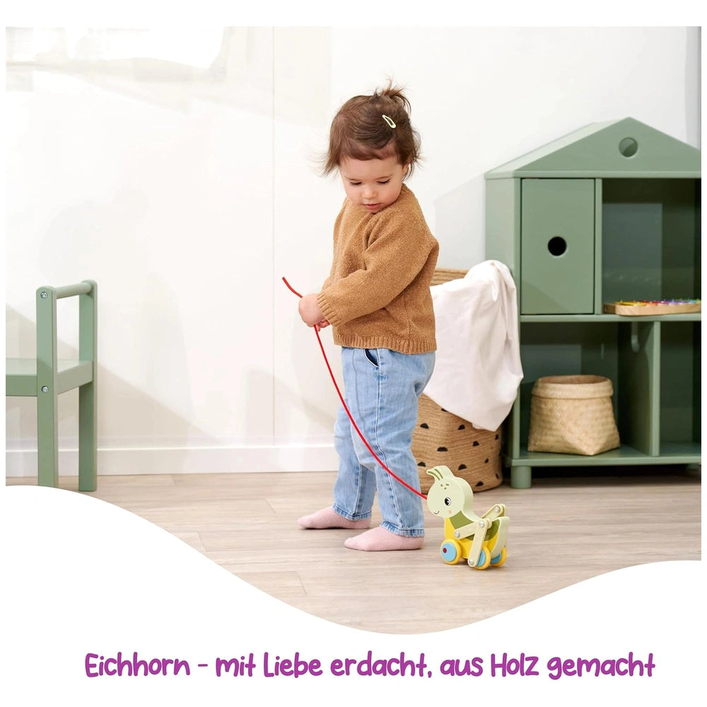 Colorful pulling grasshopper toy being held by a toddler in a cozy indoor setting with wooden floors and a green toy box.