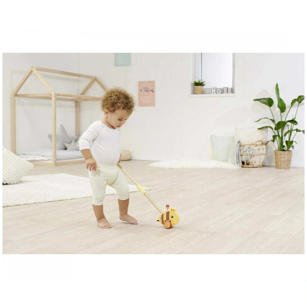 Eichhorn Push Bee toy with stick held by a child on wooden floor, bright room featuring a bed and geometric rug.