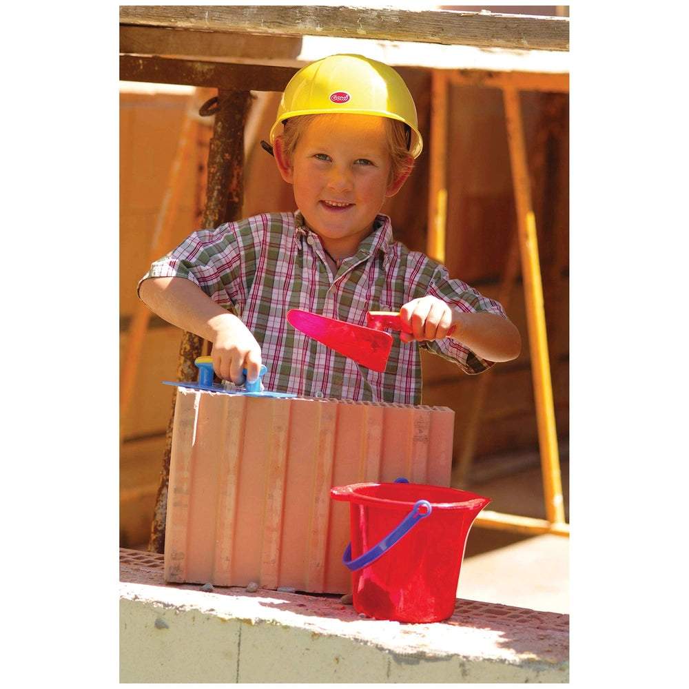Child in a yellow hard hat holds a red shovel and blue trowel, standing on gray concrete near a wooden structure and ladder.