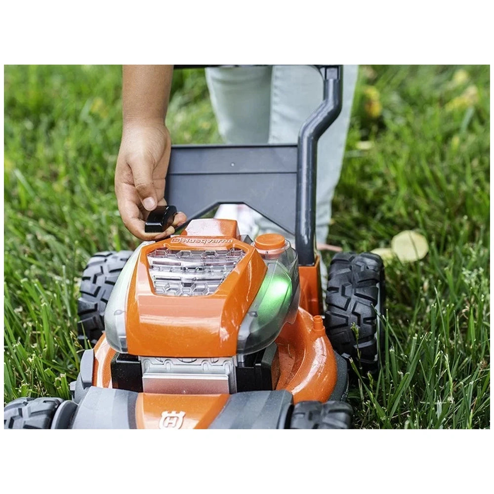 Husqvarna toy lawn mower in orange and black sits on green grass, with a person's hand wearing blue jeans visible.