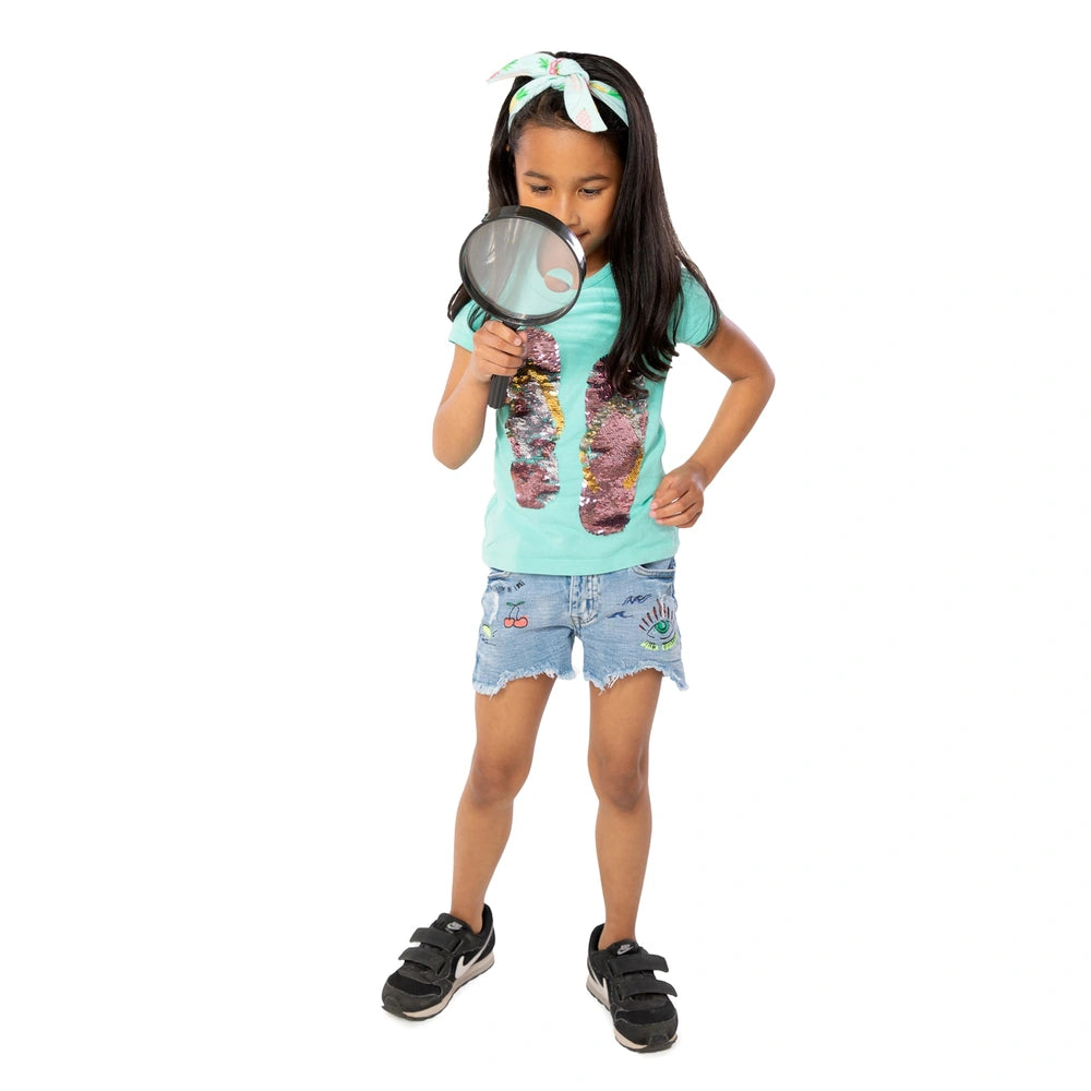 young girl with long black hair, wearing a light blue t-shirt, holding a Johntoy magnifying glass while looking down.