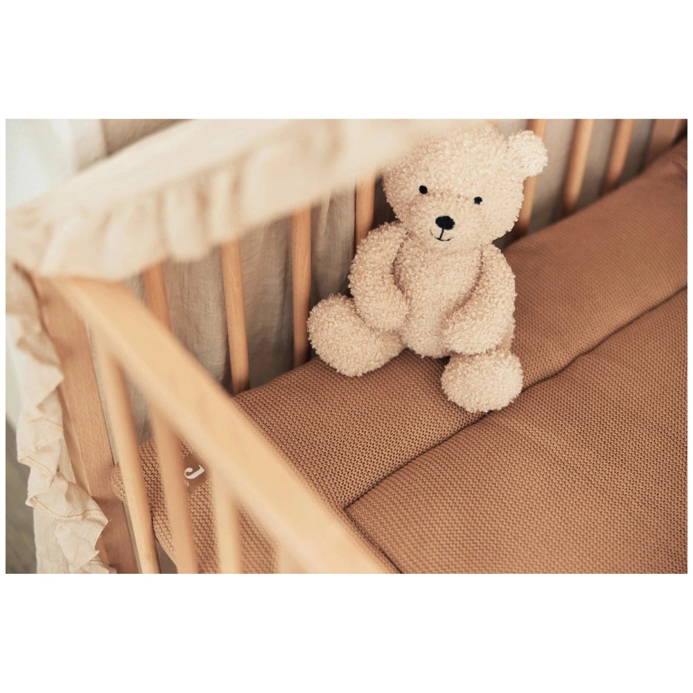 White teddy bear on light brown crib mattress inside a wooden crib frame in a cozy lifestyle scene.