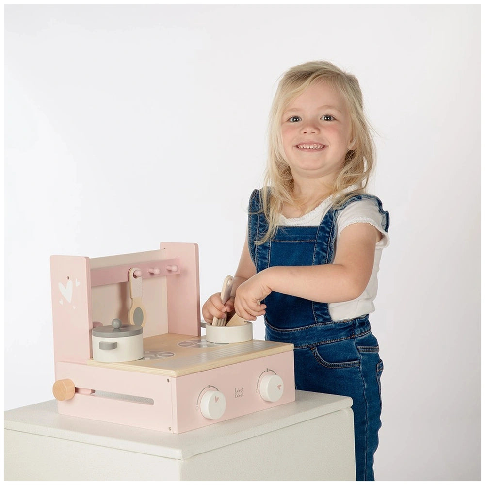 Young girl in white shirt and blue overalls smiles while holding a spoon in front of a pink toy oven with a white lid.