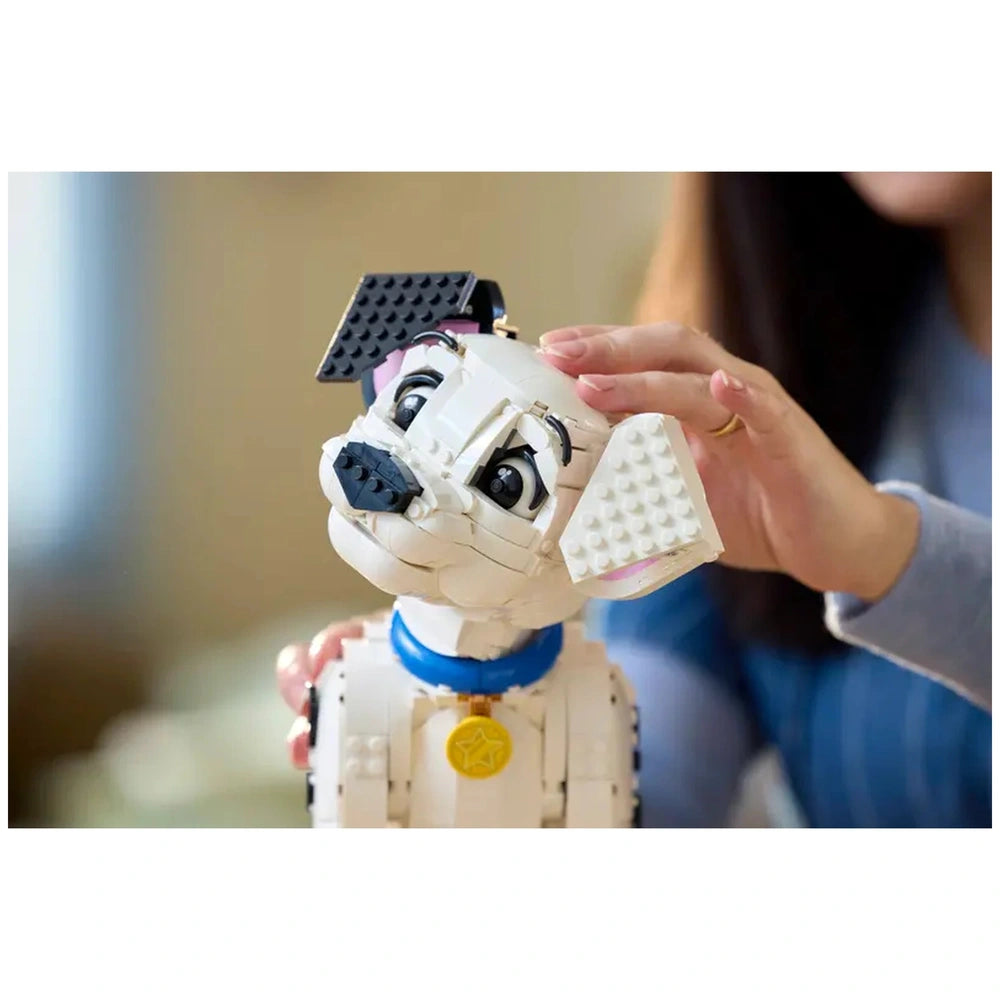 LEGO 101 Dalmatians puppy figure with a blue collar and yellow tag is being held by a person's hands in a warm indoor setting