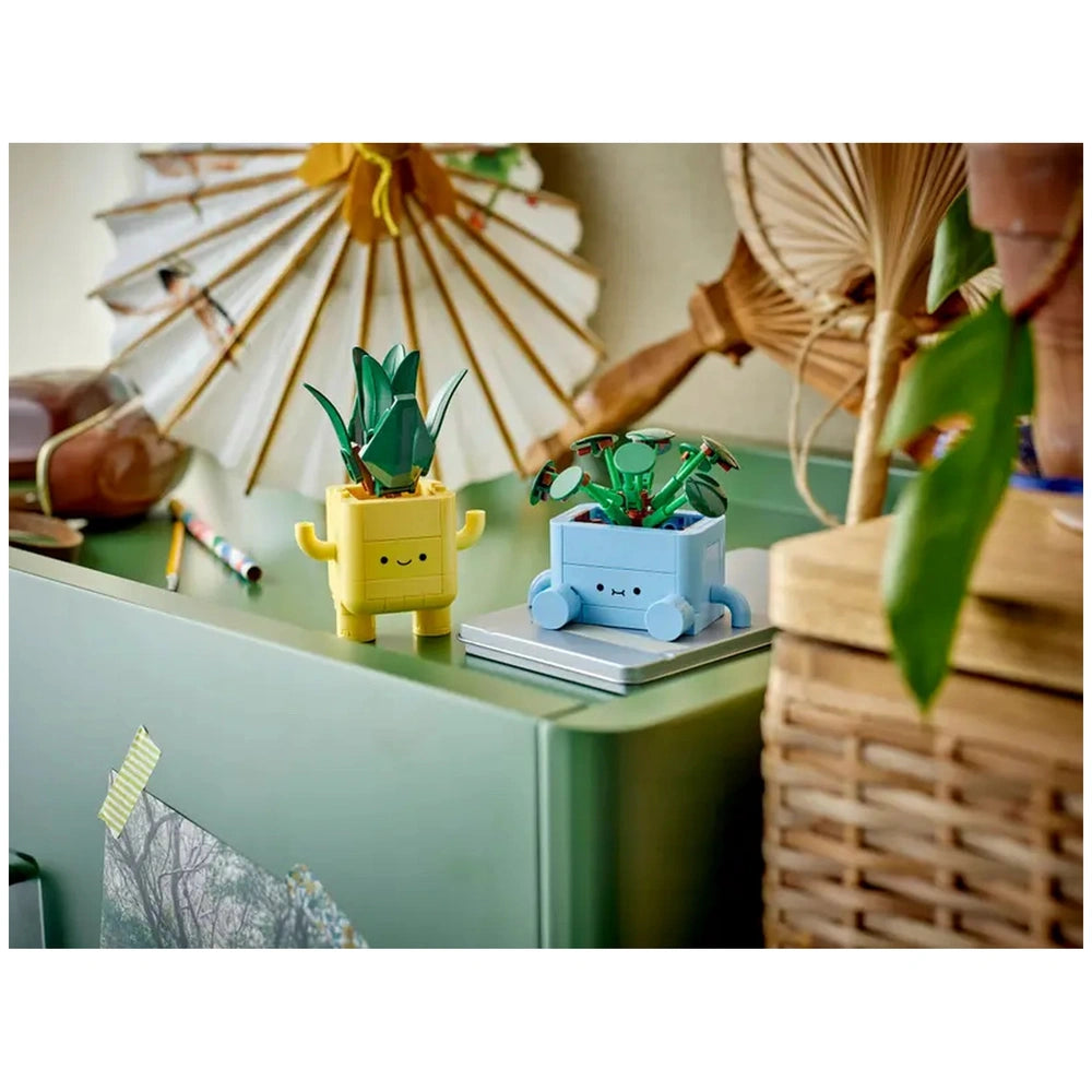 Two colorful LEGO plant pots on a green table, a yellow pot with a tall plant and a blue pot with smaller plants, surrounded by natural decor elements.