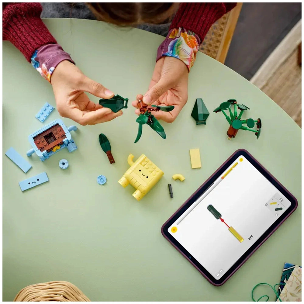 A child builds with LEGO Botanicals Happy Plants pieces on a green table, holding a plant leaf in one hand while an instructional tablet displays assembly steps. Various colorful LEGO elements and pots are scattered around.