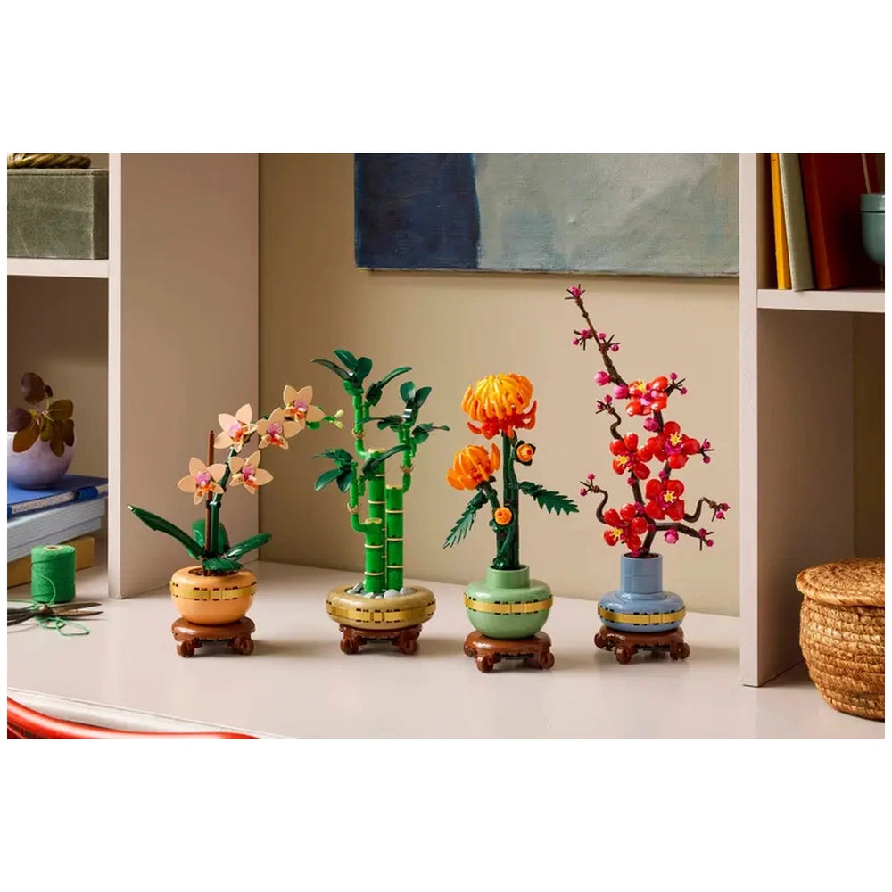 Four colorful LEGO® Botanicals plants displayed on a light surface, showcasing a pink orchid, green bamboo, a yellow flower, and a vibrant cherry blossom in decorative pots.