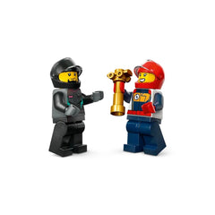 Two LEGO race driver minifigures celebrate victory, one holding a gold trophy. The first figure wears a black helmet and gray outfit, while the second dons a red helmet and colorful racing gear.