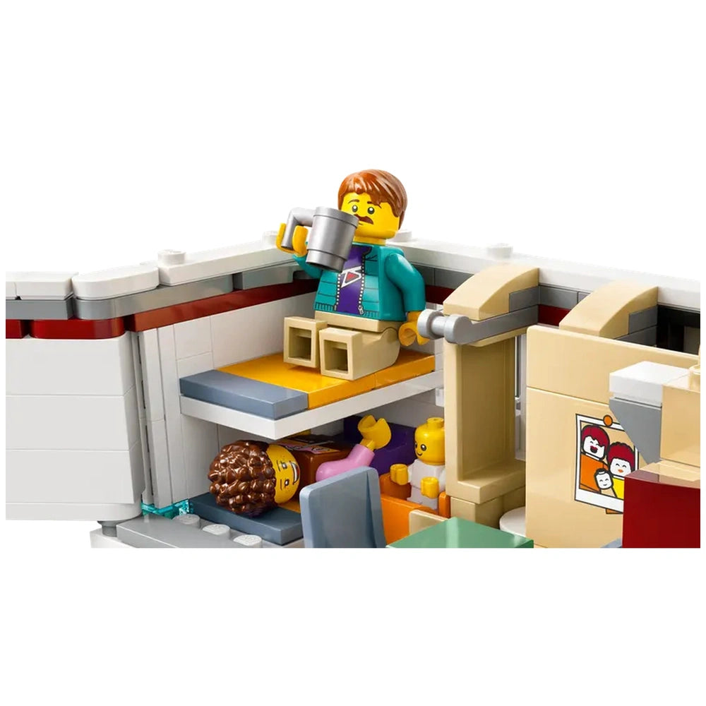 A boy in a turquoise jacket sits on the upper bunk of a LEGO camper van, holding a cup. Below, another figure lies on a lower bunk, with a child beside them, all set within a cozy interior featuring a kitchenette and colorful decor.